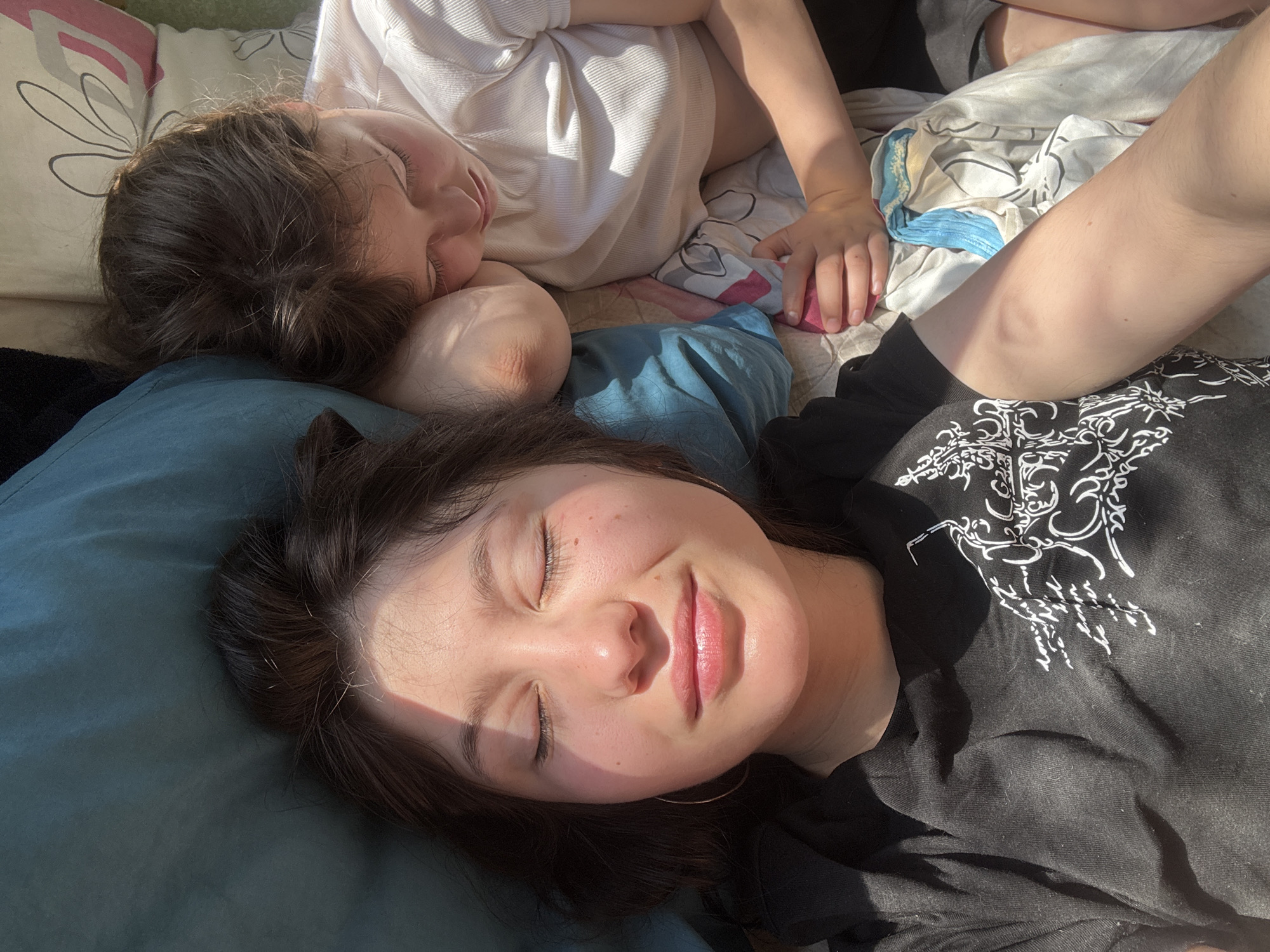 Two individuals are lying on a bed or soft surface. The person in the foreground is lying on a blue pillow. The individual in the background is nestled against a white pillow. The person in the foreground is wearing a black shirt with a white design. The person in the background is wearing a white shirt. The location is likely indoors, given the presence of bedding. There are no discernible details that confirm the location as Фоки, Russia.