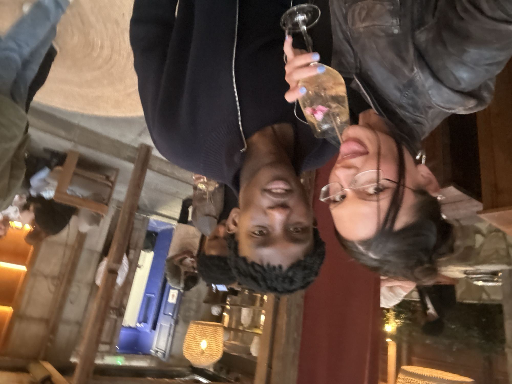 A photo shows two people holding up flutes containing a liquid with what appears to be a flower in it. One is a black man with short hair, wearing a navy blue sweater. The other is a white woman wearing glasses. She has her tongue sticking out. They are indoors in a bar/restaurant location, possibly in London.