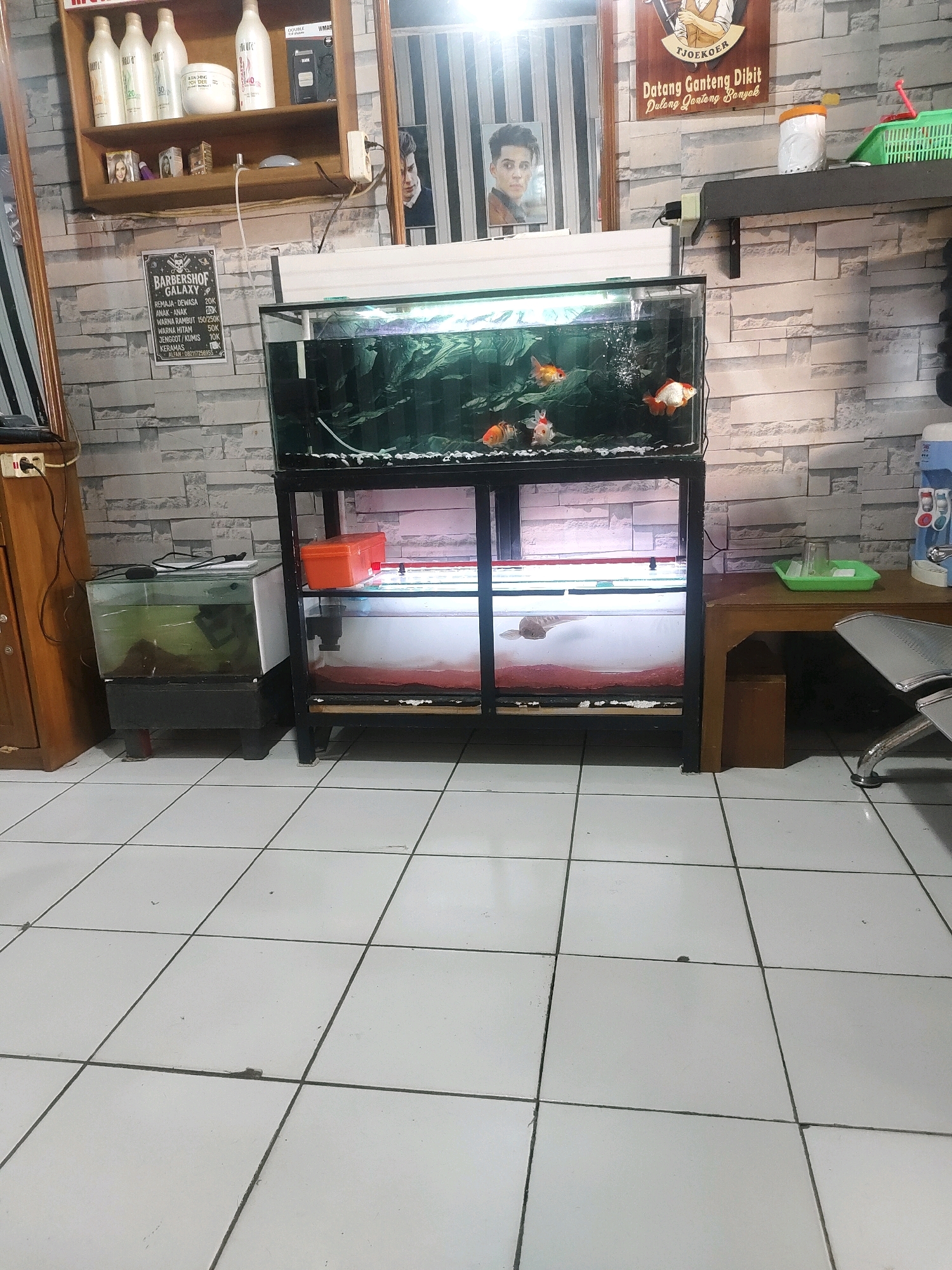 The image depicts an interior scene, likely a barbershop in Sukanagara, Indonesia, based on the presence of barber-related signage. The foreground shows white tiled flooring. In the middle ground, a large aquarium with several goldfish and at least one larger fish is prominently displayed on a black metal stand. To the left of this aquarium is a smaller glass tank with a dark substrate and some plants. To the right of the large aquarium, a wooden table holds a water dispenser, a green tray with cups, and a silver chair is partially visible. The background features a stone-patterned wall. Various items are displayed on shelves above the wall, including bottles of what appear to be hair products and framed pictures. A sign with "BARBERSHOP GALAXY" and a price list for services is also visible on the wall. The overall impression is that of a functional barbershop environment.