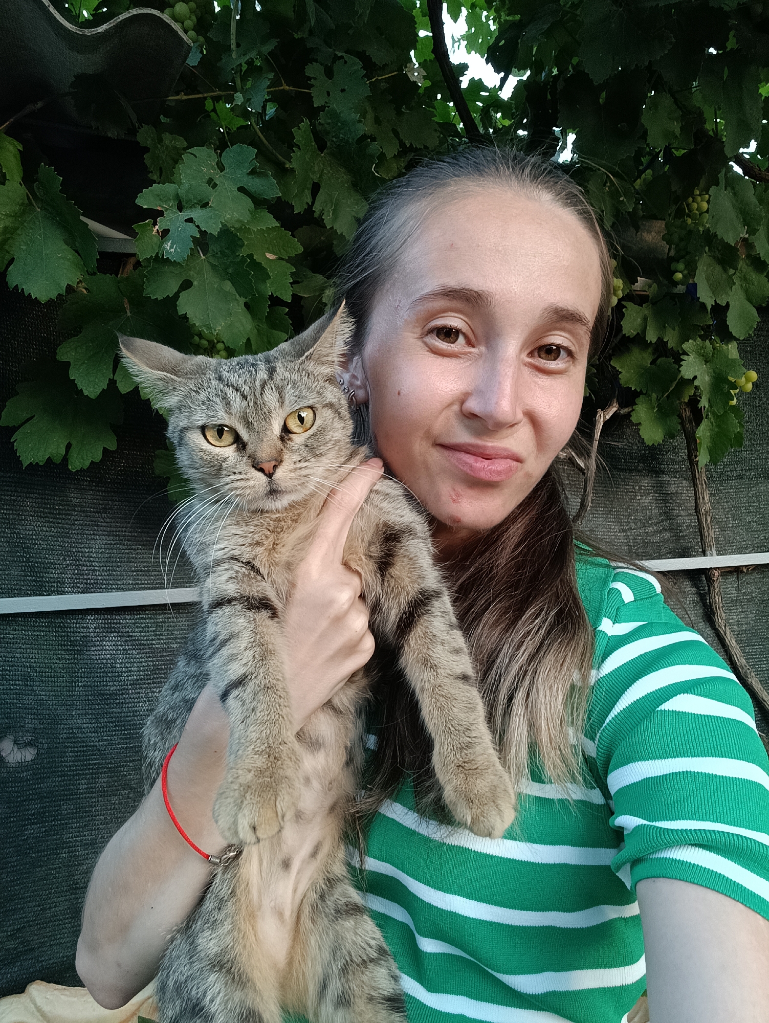 A young woman with long brown hair, wearing a green and white striped shirt, is holding a tabby cat in the city of Vypasne, Ukraine. The woman is looking at the camera with a slight smile. The cat, held in her arms, has yellow eyes and is looking at the camera with a serious expression. The background features lush green foliage, suggesting an outdoor setting, possibly a garden or park. The lighting indicates it is daytime. A red bracelet is visible on the woman's wrist. The image appears to be a selfie.