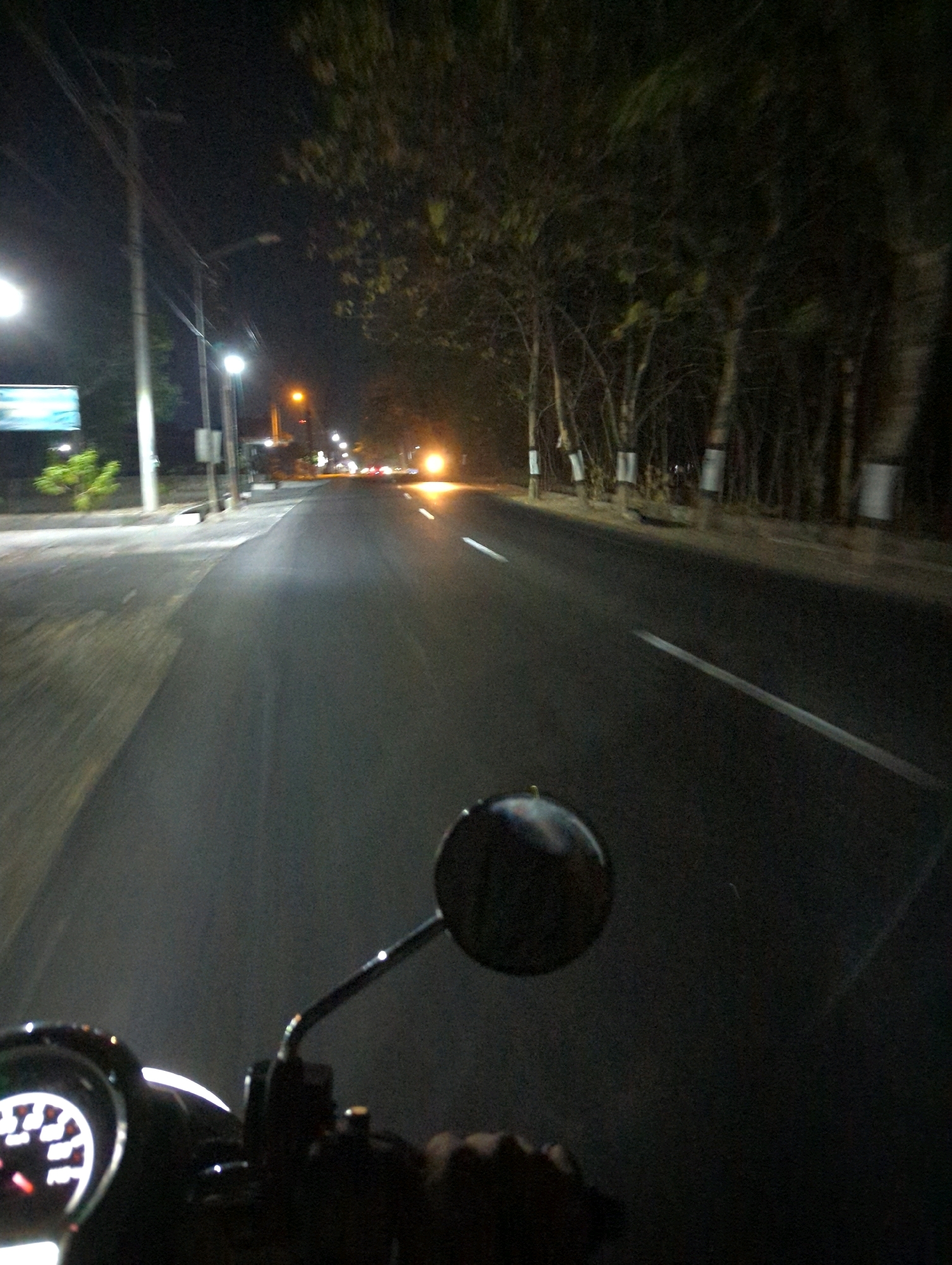 The image is a first-person perspective of someone riding a motorcycle at night on a paved road in Kacangan, Indonesia. The road is lined with trees and has streetlights illuminating sections of it. The motorcycle's dashboard, including a speedometer, and a side mirror are visible in the foreground. The overall scene depicts a typical nighttime commute or journey through a semi-urban or rural area in Indonesia.