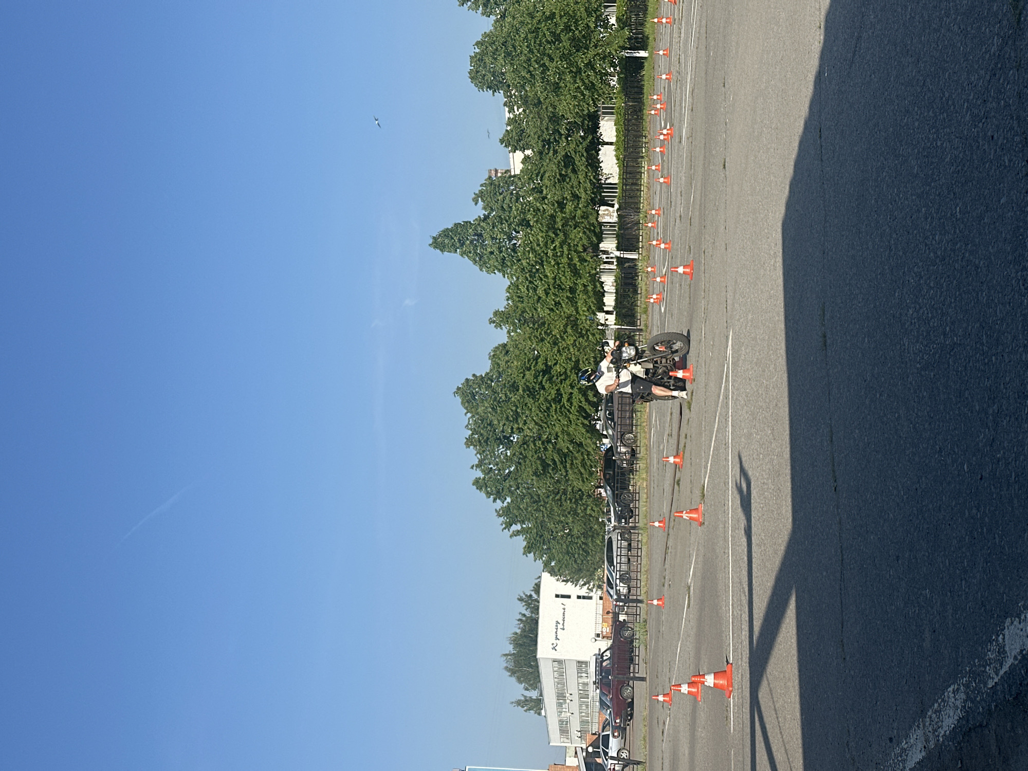The image depicts a motorcycle training area, possibly in Chaikovsky, Russia, considering the Russian text on the building. The scene is primarily an asphalt parking lot with rows of orange traffic cones. A motorcyclist dressed in protective gear is visible on a motorcycle amidst the cones. Parked vehicles line the edge of the lot, behind a metal fence. A white building with a sign in Russian sits beyond the parked cars and fence. Trees are behind the building, lining the street. The sky is clear and blue. The photo appears to have been taken during daylight, with a large shadow covering a portion of the parking lot.