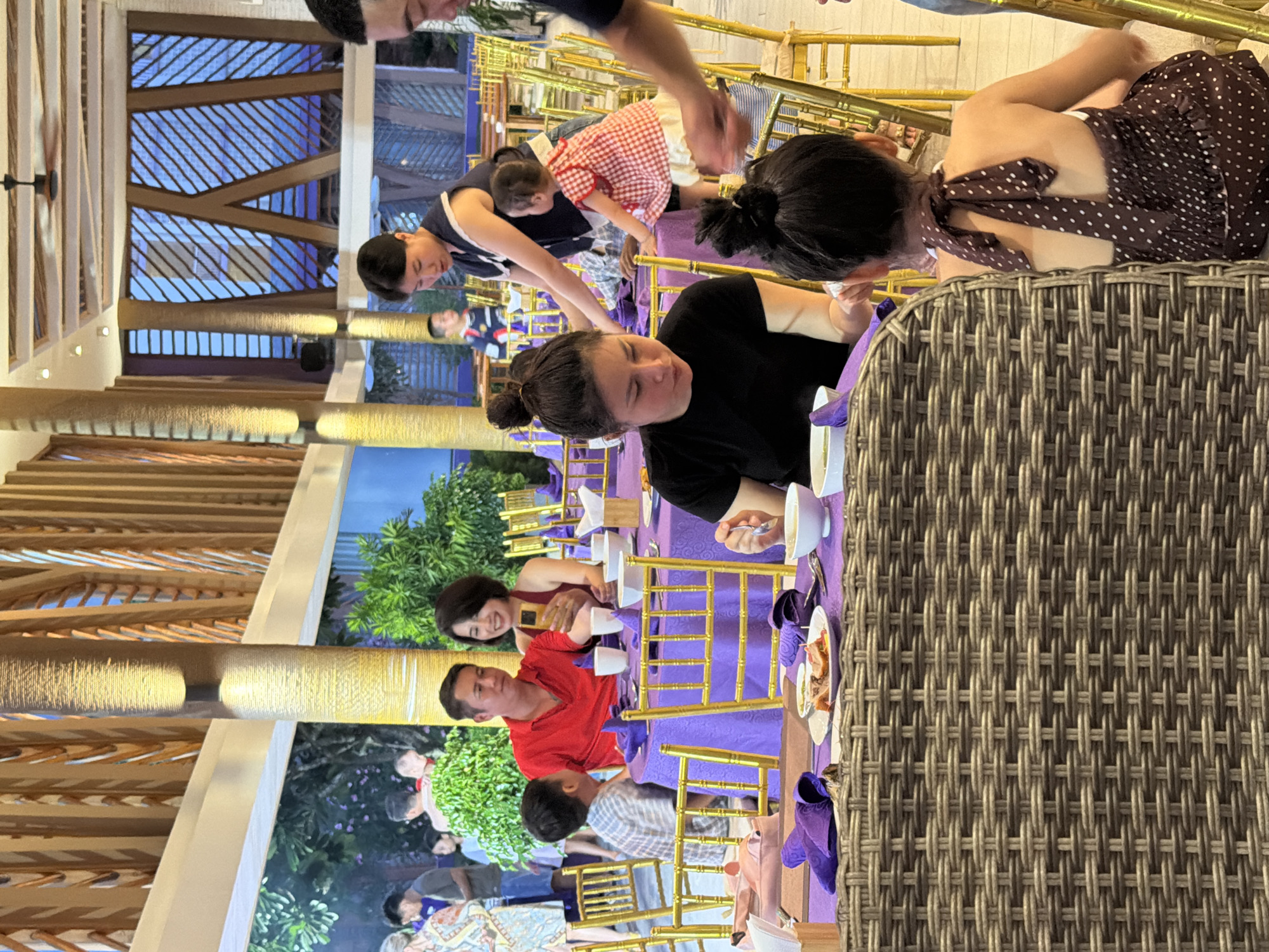 This image captures an evening dining scene within a spacious restaurant or resort dining area located in Dong Ninh Hoa Ward, Vietnam.
The environment features tall, light-colored columns with wall-mounted lights and a high ceiling with exposed wooden beams and slats. Decorative wooden lattice panels with geometric patterns are visible on the upper walls, revealing blue-tinted windows or screens beyond. Green foliage, including potted plants and shrubs, is integrated into the setting.
In the immediate foreground, the back of a woven, possibly rattan, chair is visible. A young child with dark hair in a bun is seated with her back to the camera, wearing a brown halter-neck top with white polka dots. To her left, an adult female with dark hair in a bun, wearing a black top, is eating from a white bowl with a spoon. A plate containing roasted meat and other food items rests on the purple tablecloth in front of her.
In the mid-ground, an adult male in a red polo shirt is seated at a table covered with a purple tablecloth, observing a young boy in a striped shirt seated opposite him. Next to the man in red, an adult female with shoulder-length dark hair, wearing a maroon top, is smiling and holding a smartphone, seemingly capturing a photograph or video. Further right, an adult female with short dark hair, wearing a dark top with a white collar, stands and leans over a table, attending to a small child in a red and white checkered dress standing beside her. An adult male's arm is partially visible on the far right, reaching towards the child in the polka-dot top.
The background shows several other individuals standing and seated at similar tables with golden chairs. A large blue-tinted window with a diagonal lattice pattern is prominent behind the tables on the right. A ceiling fan with wooden blades is visible in the overhead structure. White bowls, cups, and purple napkins are present on the tables.