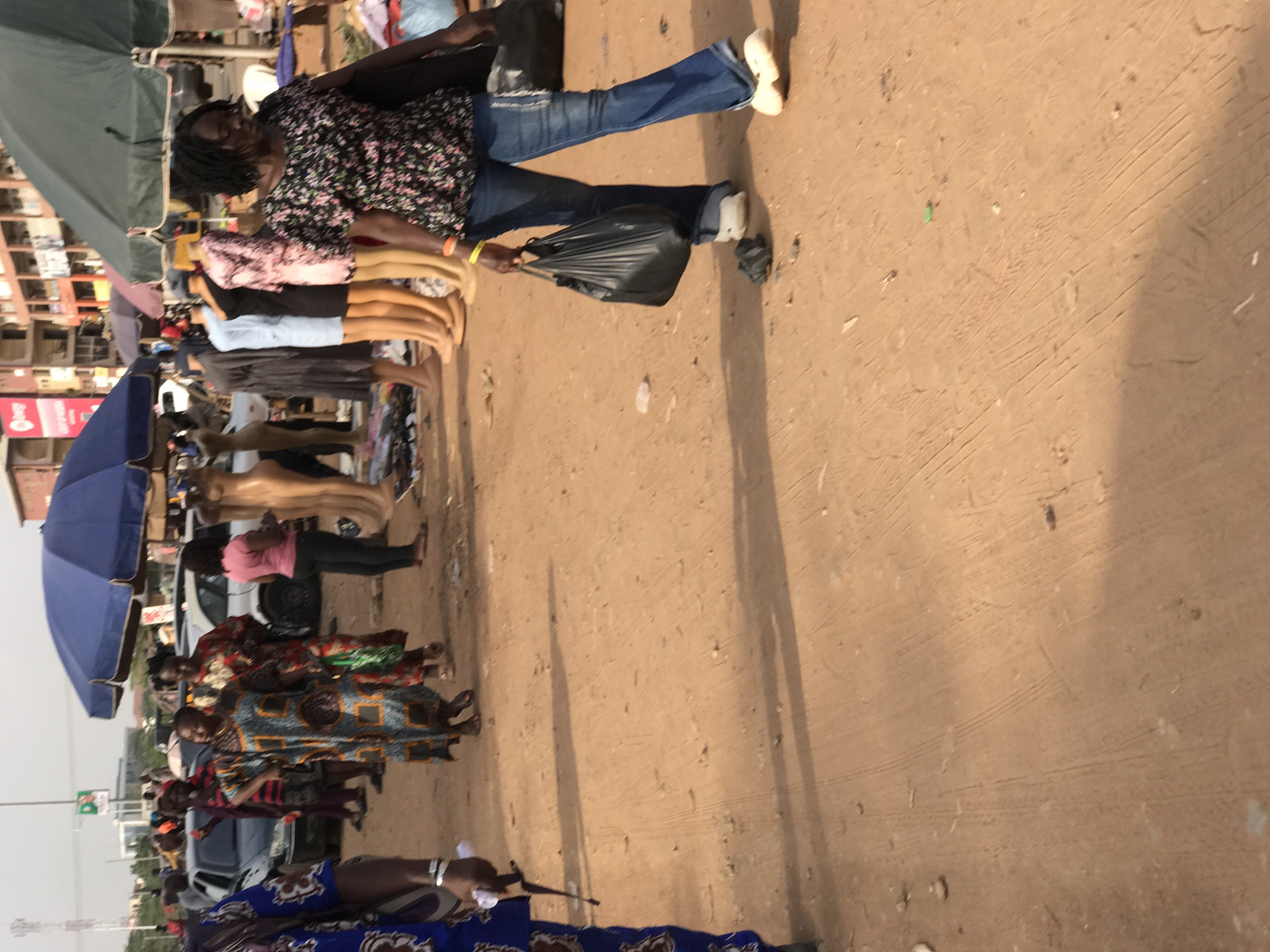 The image, originally rotated 90 degrees clockwise, depicts a bustling outdoor market scene in Abuja, Nigeria. The ground is a light brown, sandy, and unpaved surface, marked with tire tracks and footprints from heavy foot traffic.
In the mid-ground, a woman with dark hair, wearing a black floral short-sleeved top and blue jeans, walks across the frame. She carries a black plastic bag in her left hand and wears light-colored shoes. To her left, a line of mannequin legs and lower torsos, primarily light-skinned, are displayed, showcasing various dresses and tops, including a pink top, a black dress, and a light blue garment.
Further into the market, a blue market umbrella provides shade on the left, alongside a green tarp or awning that covers additional stalls. Beneath these structures, numerous people are visible, many dressed in vibrant, patterned traditional African fabrics, suggesting a cultural market setting. Other individuals are seen wearing more contemporary clothing. In the background, cars are parked or moving, indicating an urban environment.
Long, distinct shadows stretch across the dusty ground, suggesting that the sun is low in the sky, placing the time of day in either the morning or late afternoon. The lighting is bright and clear, implying good weather. On a red billboard or sign in the background, the "MTN" logo is partially visible, denoting a common telecommunications presence in the region. The scene conveys a busy, vibrant atmosphere typical of an active street market, with people engaged in commerce and daily activities.
