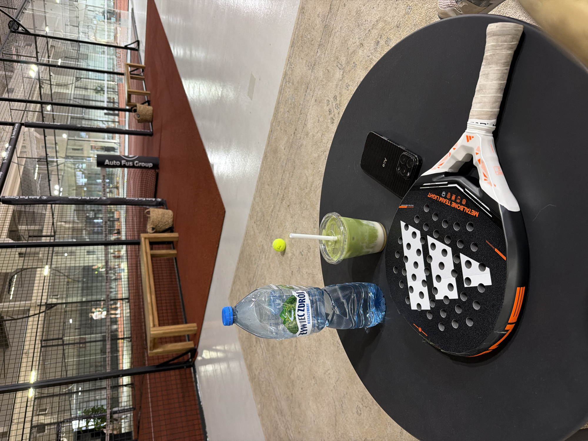 The image depicts a table with a padel racket, a phone, a drink, a water bottle, and a tennis ball on it. The table is located indoors, next to a padel court. The location appears to be in Unknown, Unknown.