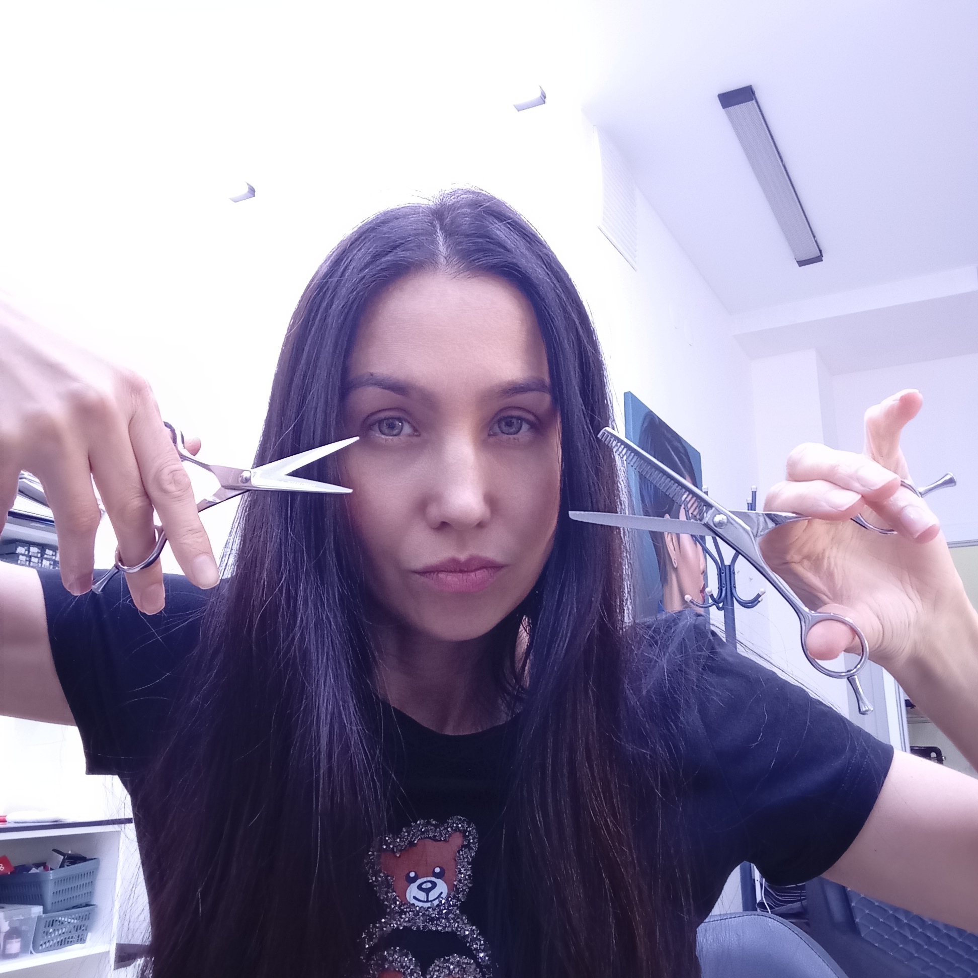 A woman with long, dark hair holds a pair of scissors in each hand, one of which has a comb, in what appears to be a hair salon. The location appears to be in Izhevsk, Russia. The woman is wearing a black t-shirt with a teddy bear design. Behind her, the salon has white walls, a fluorescent light fixture, and various salon items.