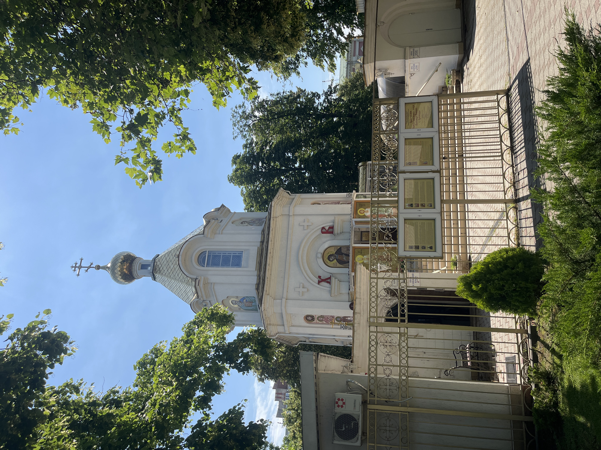 The image shows the Church of the Icon of the Mother of God "Joy of All Who Sorrow" in Krasnodar, Russia. It is a white Orthodox church with a golden dome and a cross. The church is surrounded by trees and a fence. There are several religious icons on the exterior of the church, including one depicting Jesus Christ. There are also informational boards placed outside the church. The sky is blue and clear, indicating a sunny day. The overall scene depicts a religious building in an urban setting.