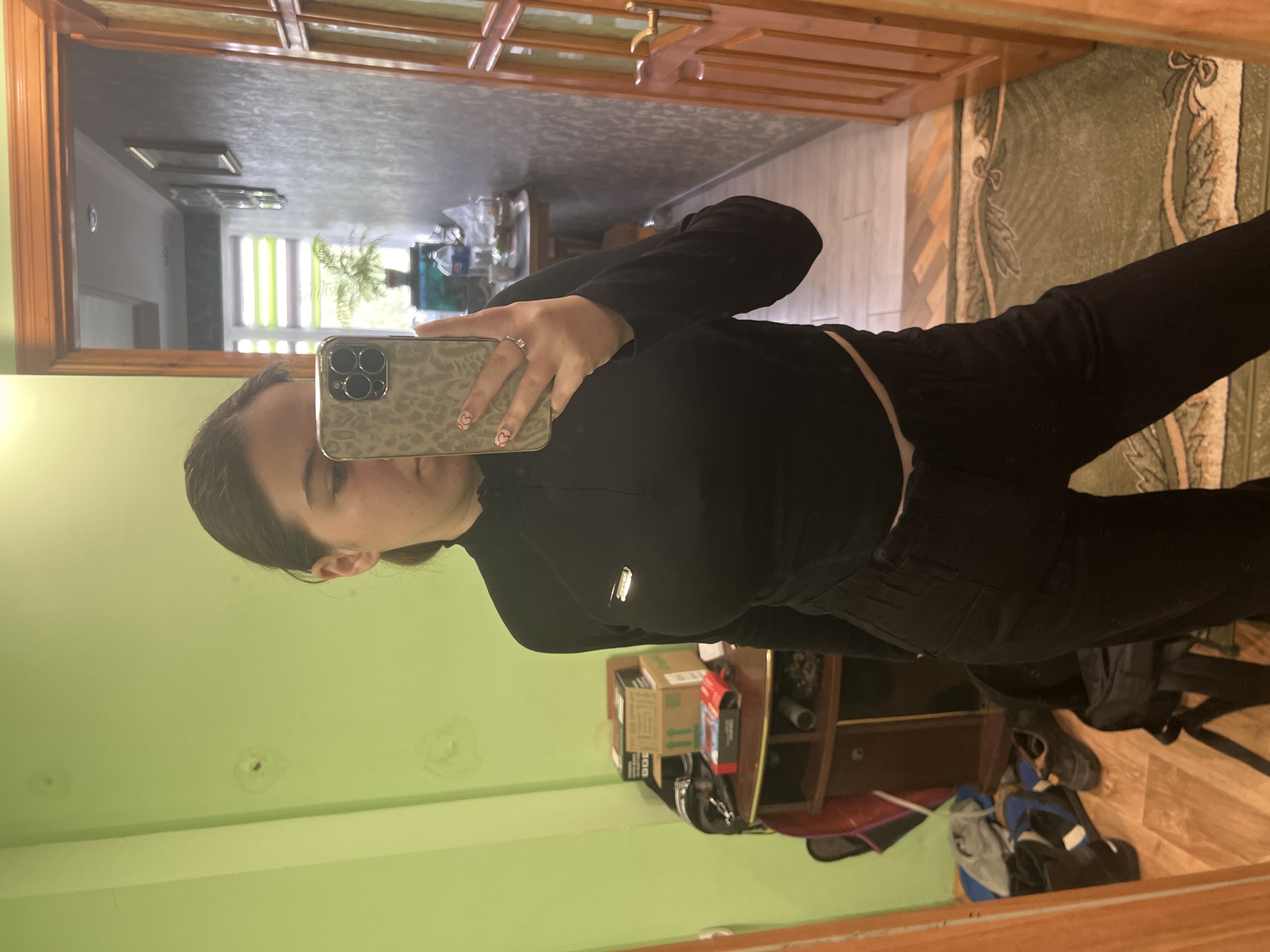 A person is taking a selfie in front of a mirror. The person has dark hair and is wearing a black long-sleeved top and black pants. They are holding a smartphone with a patterned case. The background includes a green wall, a doorway with a wooden frame, and a glimpse into another room with a window and plants. There's a cabinet with items on top of it in the reflection. The location is Cherliany, Ukraine.