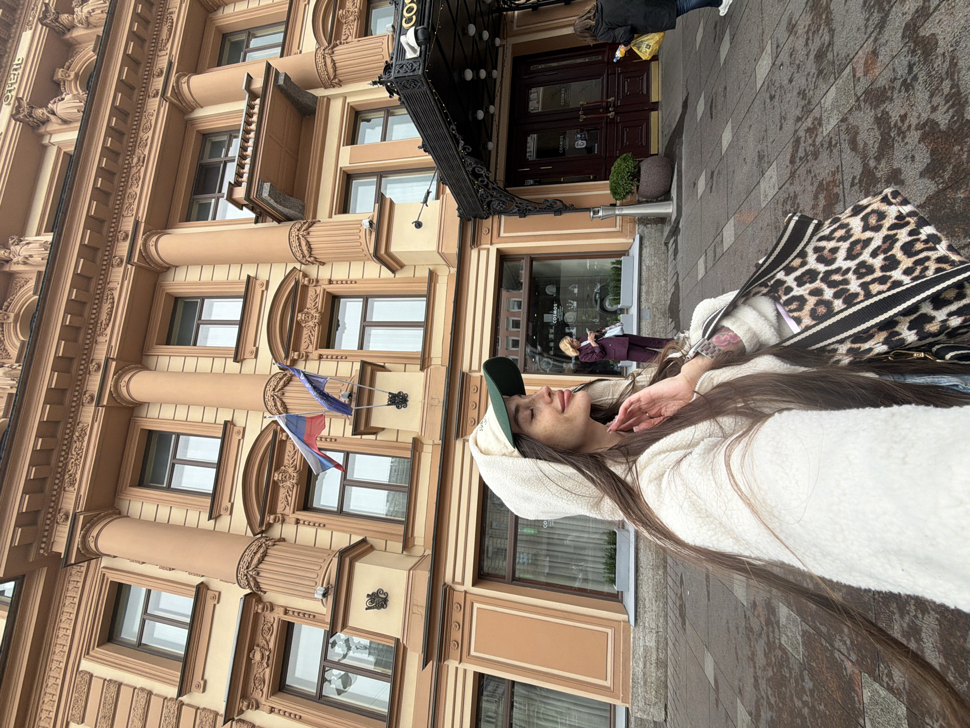 A medium shot shows a young woman taking a selfie outdoors in front of a grand, ornate building in Saint Petersburg, Russia. The building, painted in shades of beige and salmon, features classical architectural elements like columns, detailed cornices, and multiple windows. Two flags, the Russian tricolor and another blue and white flag, are visible on the facade.
The woman, facing right, is wearing a cream-colored, hooded, fuzzy jacket and a green and beige baseball cap. Her long, dark brown hair cascades down her back. She is holding a leopard-print shoulder bag with black and white striped straps in her right hand. Her left hand is raised to her face, touching her cheek. Her eyes are closed, and she appears to be smiling.
In the background, to the left, a woman in a purple suit stands on the sidewalk, looking at her phone. To the right, near the entrance of the building, a person in a dark coat is walking away, carrying a yellow bag. The building's entrance is covered by a dark awning with ornate metalwork and lighting. The pavement is wet, suggesting recent rain. The word "COSMOS" is visible on the awning, indicating the name of the establishment.