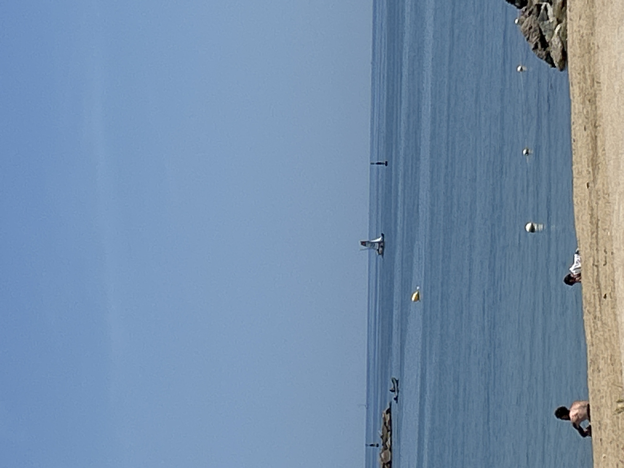 The image depicts a tranquil scene at the beach with the Mediterranean Sea stretching out under a clear blue sky. In the foreground, a sandy beach is occupied by two individuals. One person, facing away from the camera, is seated with their back exposed. To their right, another person is lying down.
The sea is calm, with gentle ripples reflecting the sky. Several buoys are visible floating on the water, indicating a marked area. In the middle ground, a small boat with two occupants is navigating the water near a rocky pier topped with a lighthouse-like structure. Further out, a sailboat with a red and white sail is gliding across the sea. On the horizon, another distant sailboat can be faintly seen. The overall impression is one of a peaceful day at the seaside, likely in a location within Barcelona, Spain, given the context provided.