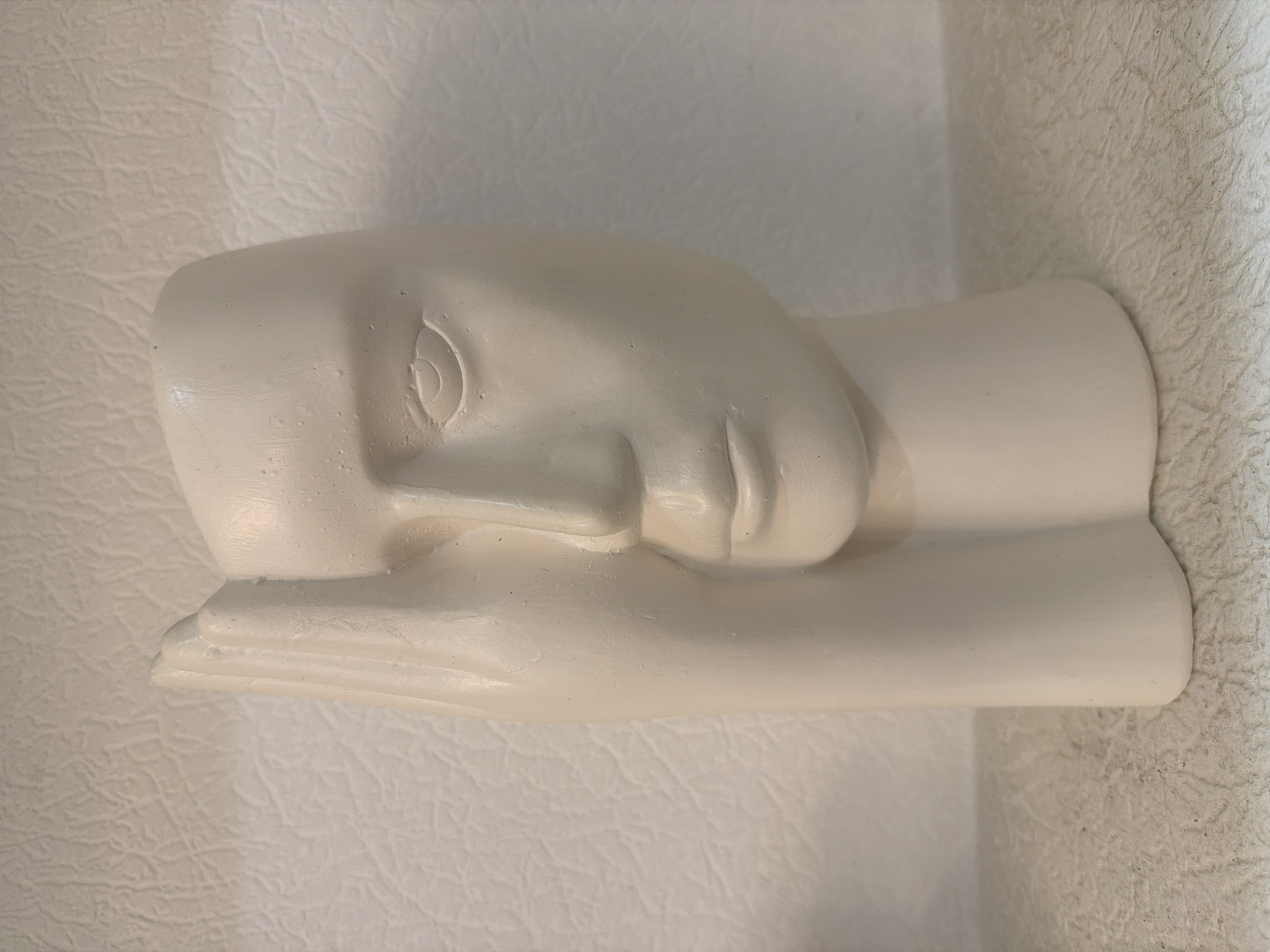 A matte white ceramic vase in the shape of a human face, with a hand covering the left side of the face and the eye. The vase has subtle facial features like an eye, nose, and mouth, and its base is a rounded, pedestal-like form. The background is a textured, off-white surface, possibly a wall. The vase sits on a similar textured surface, likely a shelf or table. The image is taken in a well-lit indoor environment. The location is not specified.