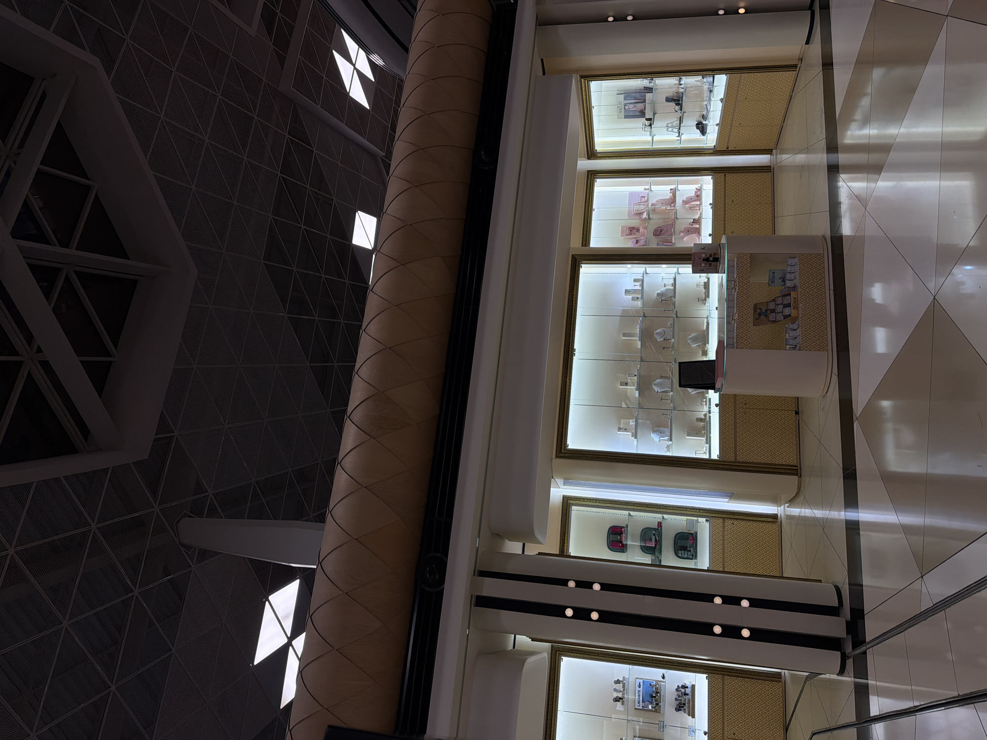 The image depicts the interior of a brightly lit retail establishment, likely a jewelry or accessories store, situated in Khazar Raion, Azerbaijan. The store features multiple display cases with glass fronts, showcasing various items on shelves. The display cases are illuminated from within, highlighting the merchandise. One display case on the left exhibits watches and a Lacoste branded item. Another case in the center displays necklaces and bracelets, with some pink packaging. A third case on the right also contains watches and other accessories.
In the foreground, there is a curved counter with a display of small packaged items, some of which have text on them. A tablet or screen is visible on the counter. The floor is a polished surface with a geometric pattern of light and dark tiles.
Above the display cases, a decorative, curved element runs horizontally across the upper portion of the storefront. The ceiling is visible in the background, featuring a dark, geometric grid structure with some illuminated panels or skylights. The overall environment suggests a modern and upscale retail setting. There are no individuals visible in the frame.