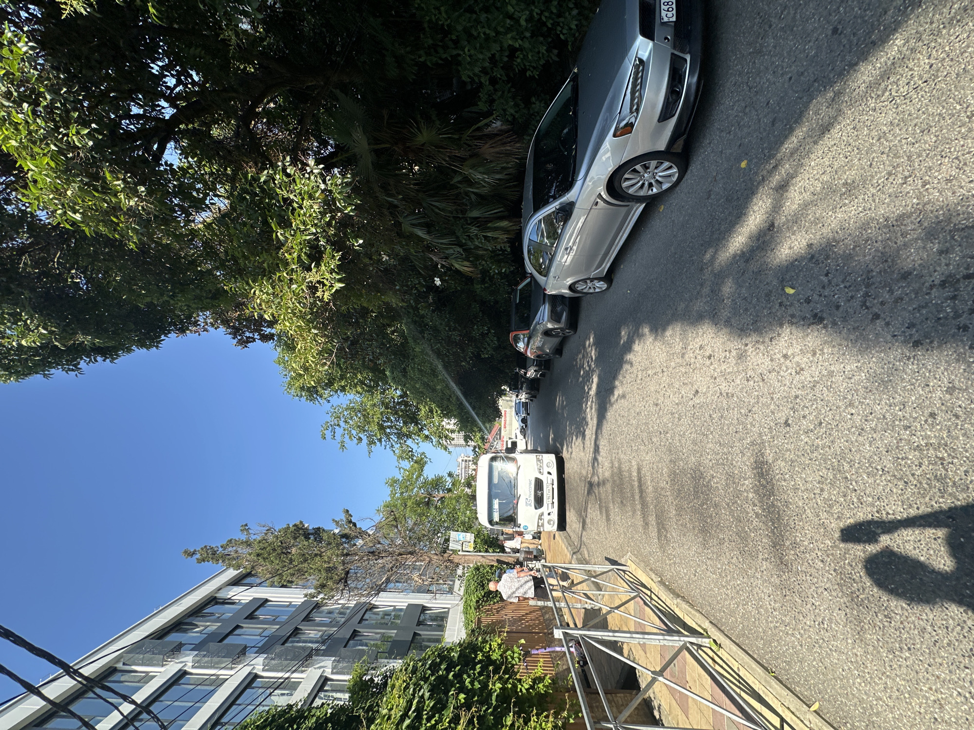 In Sochi, Russia, a paved street lined with trees and a modern building on the left is depicted under a clear blue sky. A white bus is stopped on the right side of the street, while several cars are parked further down the road. On the sidewalk to the left of the bus, a man in a light-colored shirt and dark pants walks away from the camera. The street and sidewalk are sunlit, casting shadows from the trees and a person in the foreground. A metal fence separates the sidewalk from a paved area with a pattern of yellow and grey tiles.