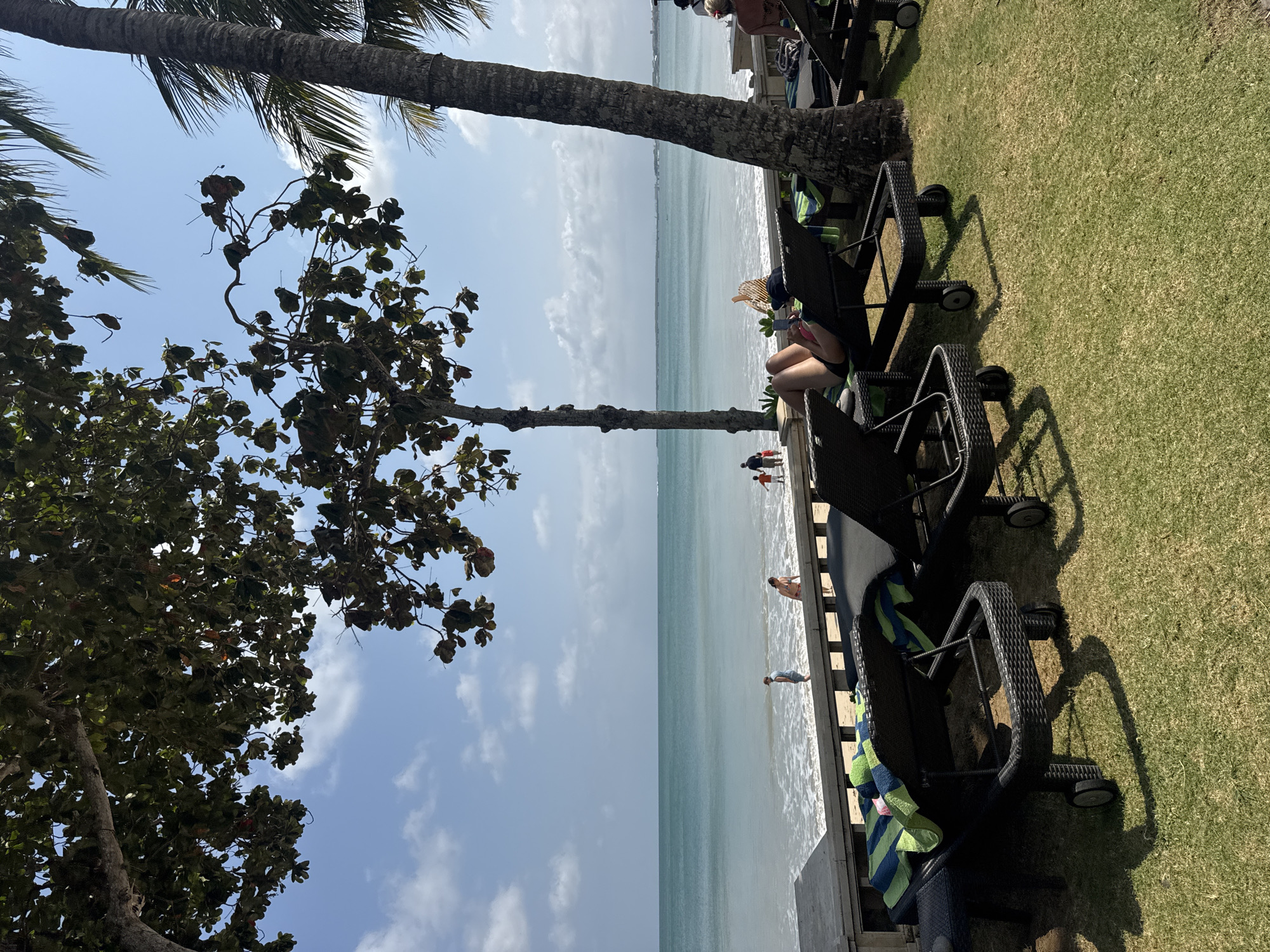 In Kuta Selatan, Indonesia, a tranquil beach scene unfolds under a bright, partly cloudy sky. The turquoise ocean laps gently onto a sandy shore, dotted with a few people walking near the water's edge. In the foreground, several dark-colored lounge chairs are arranged on a neatly manicured green lawn. One person is reclined on a chair, facing away from the camera, enjoying the seaside ambiance. Palm trees and leafy trees provide shade, casting dappled shadows on the grass. The scene evokes a sense of relaxation and a perfect day at the beach.