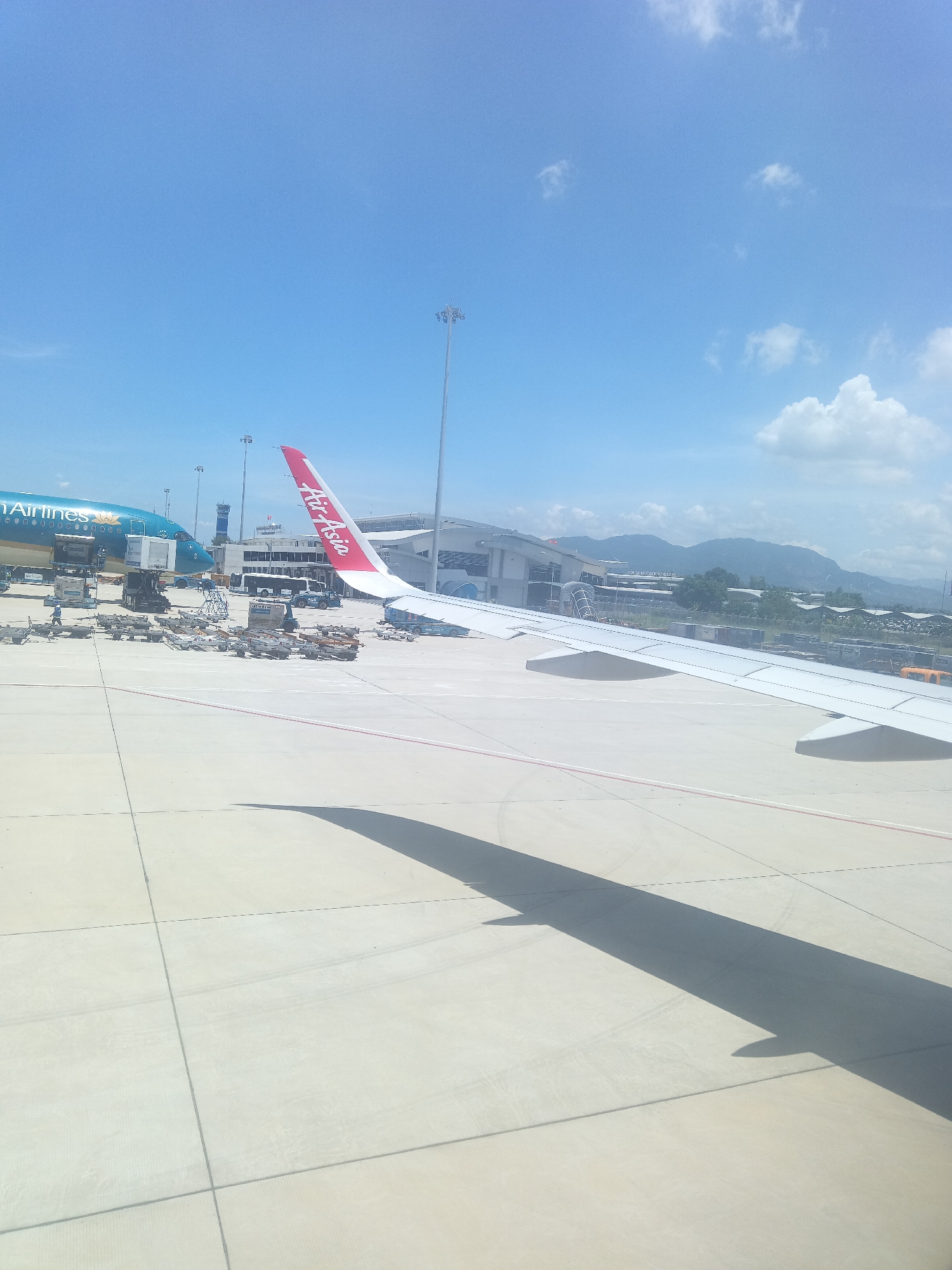 This image captures an airport tarmac scene in Bắc Cam Ranh, Vietnam. In the foreground, a section of a white airplane wing extends across the frame, casting a long shadow on the concrete surface. The wing is attached to an airplane with a bright red tail fin featuring the "Air Asia" logo in white lettering. To the left, another airplane, predominantly teal with "Vietnam Airlines" branding, is parked near a jet bridge. The tarmac is marked with grid lines and a red stripe running parallel to the wing. Various airport equipment, including baggage carts, loaders, and a bus, are visible around the parked aircraft. In the background, airport buildings and terminals are situated, with a mountainous landscape and a clear blue sky with scattered clouds forming the backdrop. The overall scene depicts a typical operational environment at an airport on a sunny day.
