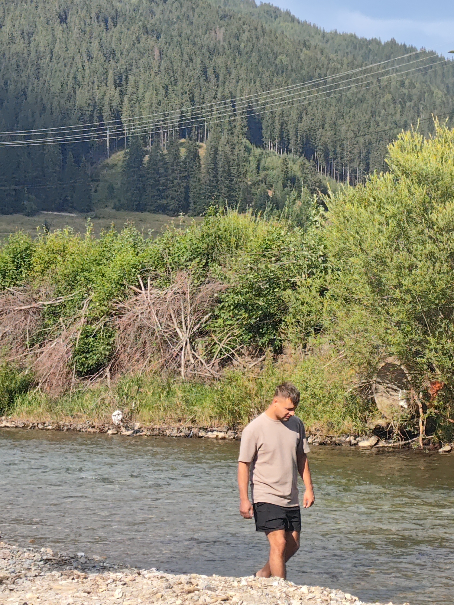 A young man wearing a beige t-shirt and black shorts is standing in a shallow, clear river. He is looking down and appears to be walking through the water. The riverbank is lined with rocks and green vegetation, with dead branches and debris scattered near the water. In the background, a dense forest covers a hillside, with power lines running across it. The sky is clear and blue. The scene is set in a natural environment, likely a rural or mountainous area. The image does not provide specific details about the city of Botoș, Romania.