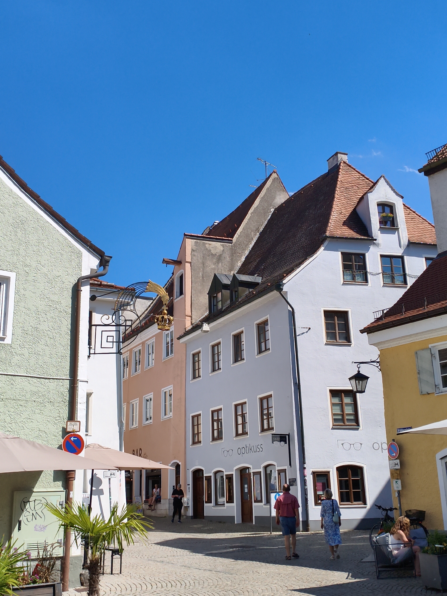 This image captures a sunny day in Landsberg am Lech, Germany, specifically on the street known as "Hintere Hölzern." The scene is dominated by several colorful, historic buildings with traditional tiled roofs. On the left, a light green building has a sign with a crown and a golden eagle. Adjacent to it, a light orange building displays "BAR CAFE" above its entrance. Further to the right, a pale blue building features the sign "optikuss" with stylized glasses, indicating an optician's shop. The street itself is paved with cobblestones. In the foreground, there are two large beige umbrellas providing shade over outdoor seating areas. Several people are visible: a man and a woman walking away from the camera on the cobblestone street, and a group of people sitting on a bench on the right side of the image, near a yellow building. A bicycle is parked near the bench. The sky is a clear, vibrant blue, suggesting pleasant weather.