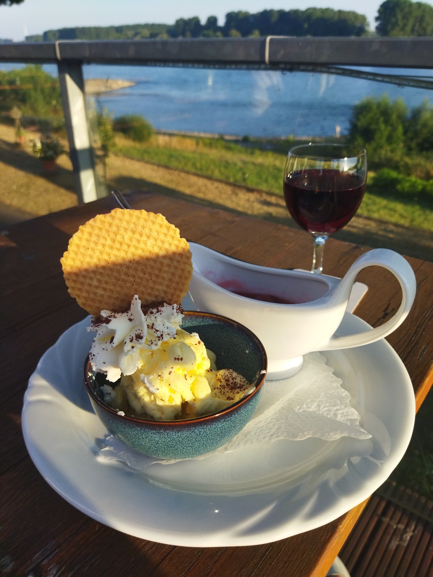 A dessert of ice cream with whipped cream and a waffle, served with a side of berry sauce in a white gravy boat, is placed on a white plate on a wooden table. In the background, a glass of red wine sits near the gravy boat, and beyond that, a body of water is visible, lined with trees and greenery. A metal railing with supporting pillars is present in the foreground. The scene is illuminated by sunlight, casting shadows. This image was taken at the Rheinpark in Monheim am Rhein, Germany.