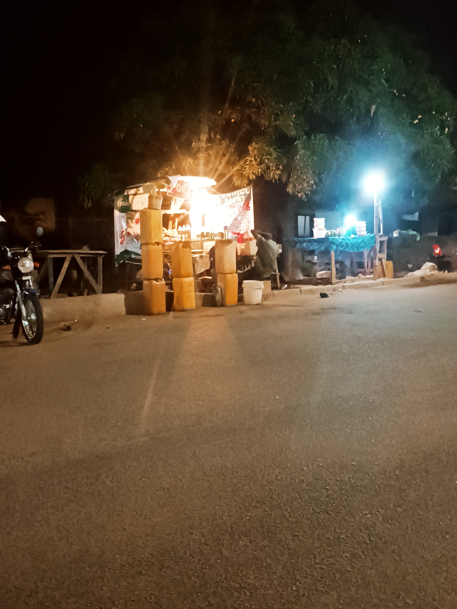 At night in an unknown city, a street market is active, lit by bright lights. A motorbike sits to the left of the frame, while stacked jerrycans stand near a market stall. The stall seems to sell various items, attended by at least one person. The scene is framed by a large tree overhead. The street is dark and empty save for the activity at the market.