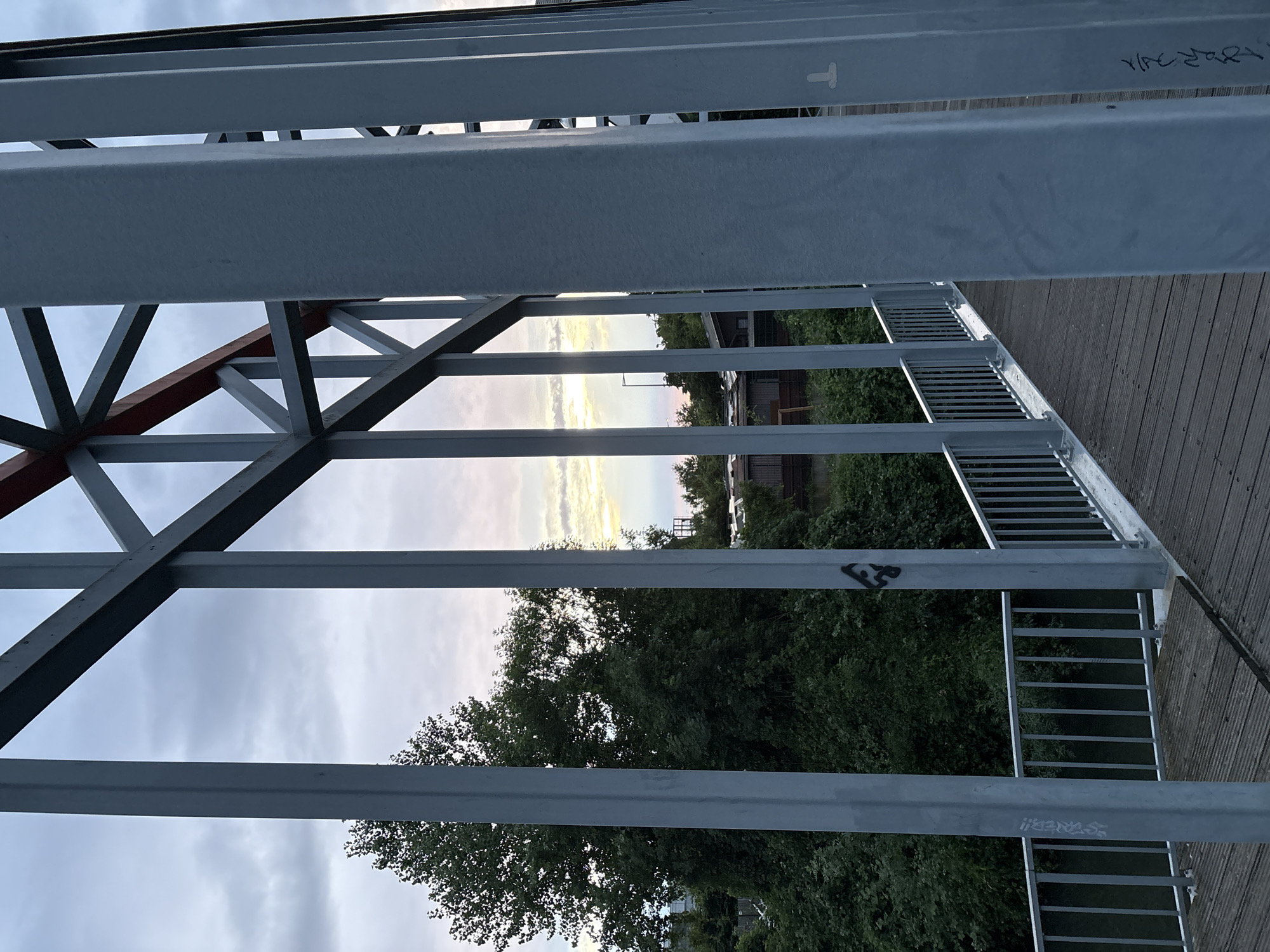 The image shows a view from a metal structure, likely a bridge or elevated walkway. The perspective is looking outwards through the metal beams and railings. In the distance, trees and some buildings are visible. The sky is cloudy with a hint of sunset colors. There is graffiti on some of the metal beams. The location and specific city are not identifiable from the image.