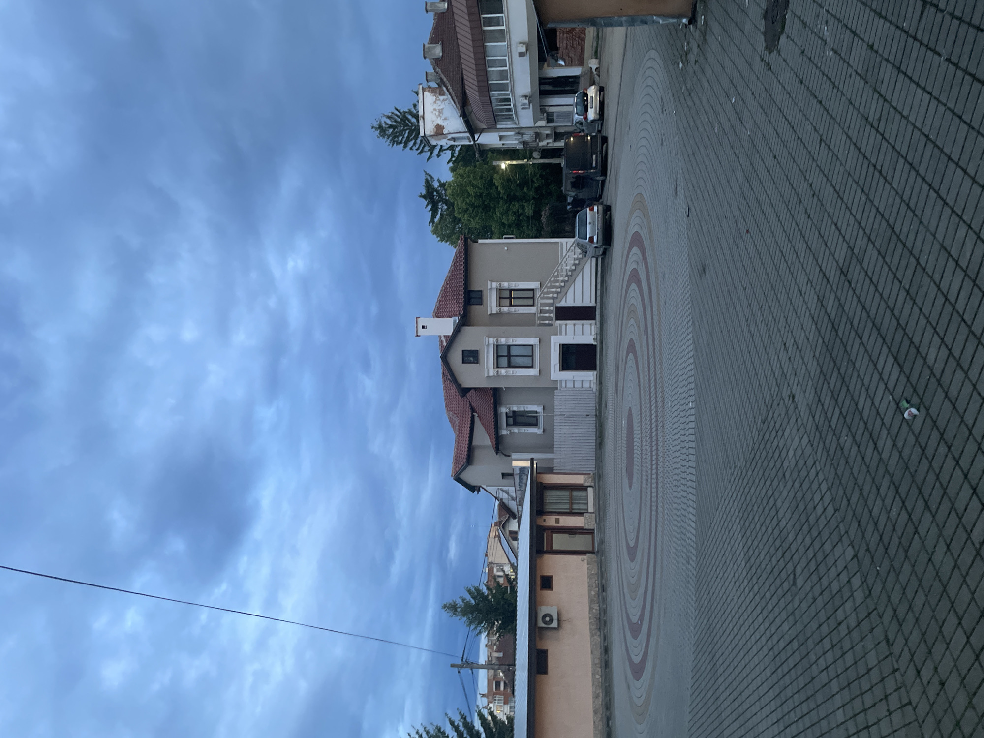 This image captures a street view in Ihtiman, Bulgaria, under an overcast, light blue sky, suggesting late afternoon or early evening light. The scene is quiet, with no human activity visible.
In the mid-ground, a prominent, well-maintained two-story house with beige walls and a red-tiled roof stands out. It features white window frames and a distinctive external staircase with white balusters leading to its main entrance. To the left of this house, part of another building with a similar red roof is visible. Further into the background, a variety of other buildings can be seen, including one with an aged, white facade and another with more modern elements. Green trees are scattered throughout the scene, nestled between and behind the buildings.
The foreground and right side of the image are dominated by a paved street or pathway. The paving consists of dark grey rectangular blocks arranged in a grid pattern, alongside a decorative section featuring concentric circles in lighter shades of beige and red. Several cars are parked along the street further down, including a white sedan and a dark SUV. Overhead, a thin power line spans across the sky. An air conditioning unit is visible on the exterior of a lower building towards the left.
The architecture, particularly the tiled roofs, suggests a European setting consistent with Bulgaria. No discernible text is visible anywhere in the image.
