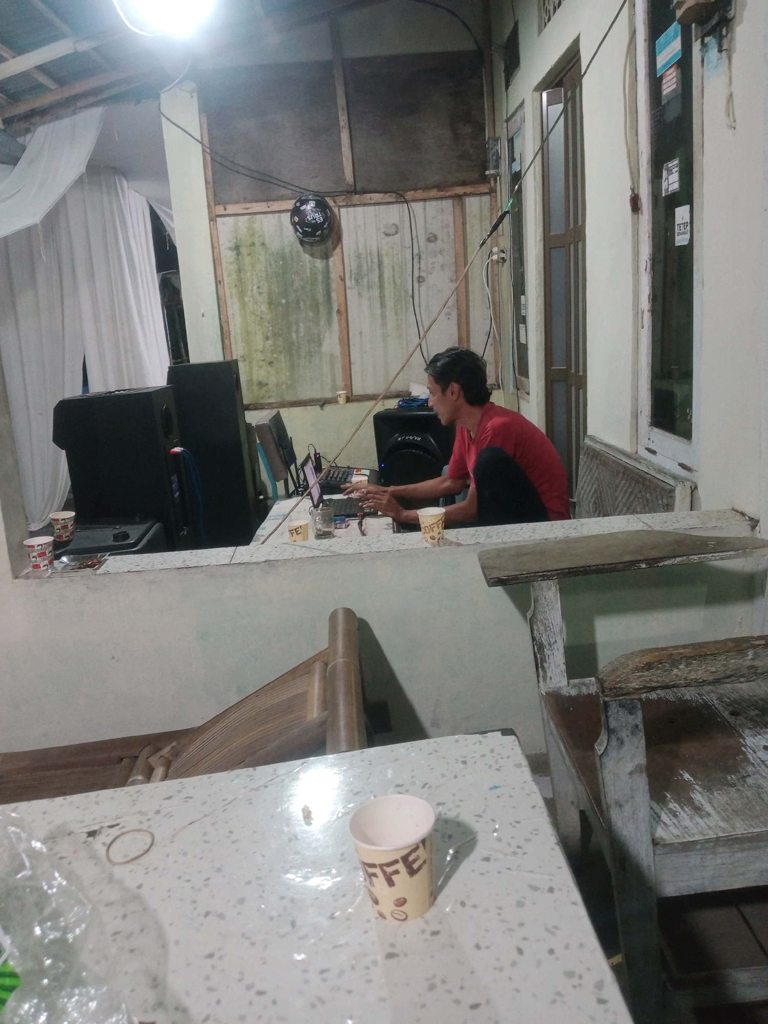 In the city of Rejasari, Indonesia, a man in a red t-shirt and dark pants is seated at a counter, engrossed in using a laptop. The room appears to be a casual workspace, possibly a home office or a small business setup. Behind the man, a large black speaker is visible, suggesting a connection to audio equipment. The counter itself is cluttered with several items, including cups, a small plate with what appears to be food, and a tablet. The environment is dimly lit, with a bright overhead light illuminating the scene. The walls are a mixture of light-colored paint and exposed wooden planks, some of which show signs of wear and moss growth. A window with a sliding door and a partial view of another room is to the right. Outside the window, a wooden bench is visible. In the foreground, a portion of a speckled countertop is present, with another cup and some plastic wrapping. The overall impression is of an informal and possibly slightly neglected space, where work is being done amidst everyday objects.