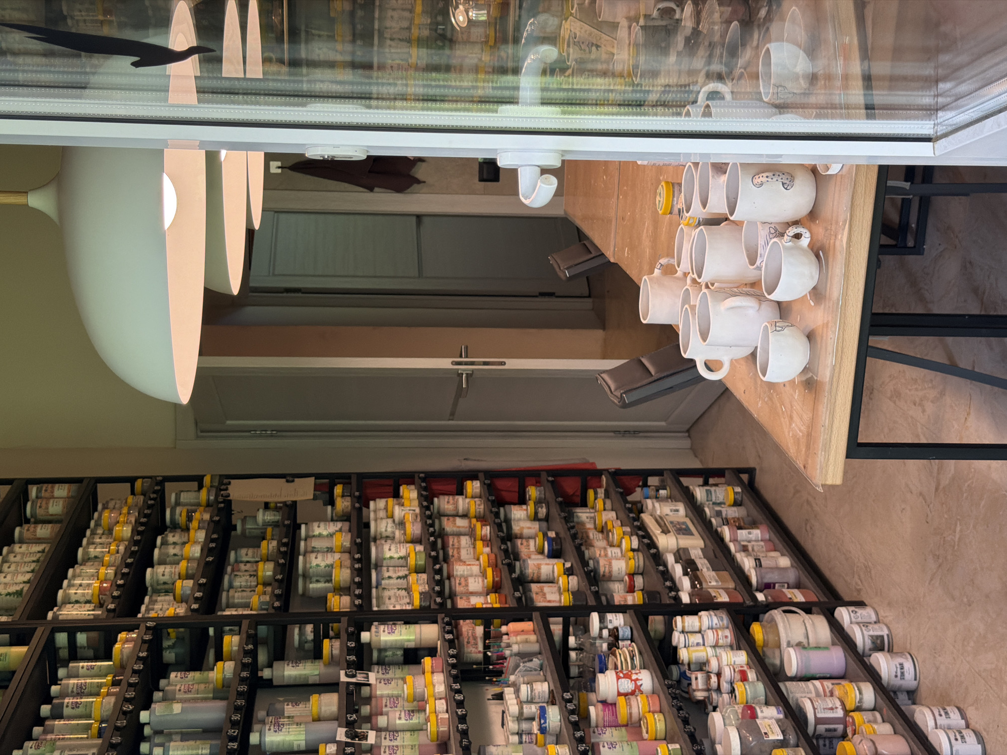 The image depicts a studio or workshop space with shelves filled with ceramic materials, likely glazes or pigments, organized in numerous small containers. The shelves are tall and appear to be made of dark metal. In the foreground, a wooden table is set with several unglazed ceramic cups and bowls, some of which have simple drawings or markings. The environment suggests a place for pottery creation or production. The location is specified as Askat, Russia. There are no people visible in the image. The lighting is artificial, emanating from overhead fixtures, and there is a large window or glass door on the right side of the frame, which reflects the interior of the room.