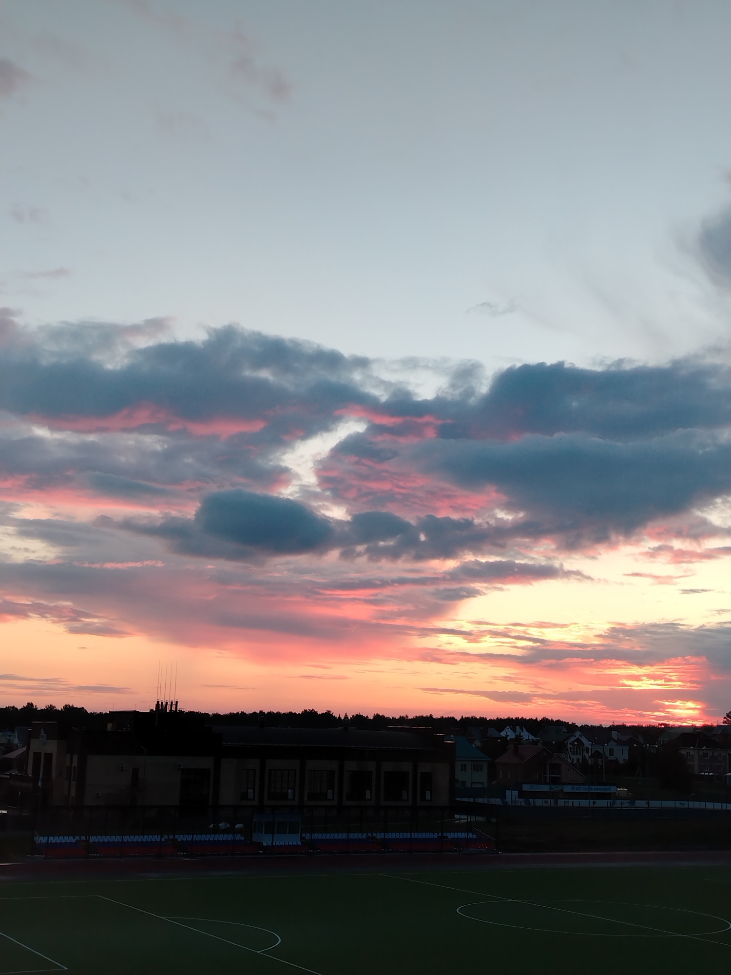 The image captures a sunset over a suburban area in Noviy, Russia, likely a residential district adjacent to a sports complex. The sky is dominated by dramatic clouds tinged with shades of pink, orange, and gray, indicating the sun is low on the horizon. Below the sky, silhouetted against the colorful sunset, are numerous residential houses with varied rooflines and some with visible windows. A long, dark building, possibly a stadium or a community center, runs along the bottom of the image, featuring a row of stadium seating in front. In the foreground, a section of a green sports field with white markings is visible, suggesting a park or recreational area. The scene is peaceful and depicts the end of a day in a moderately developed area.