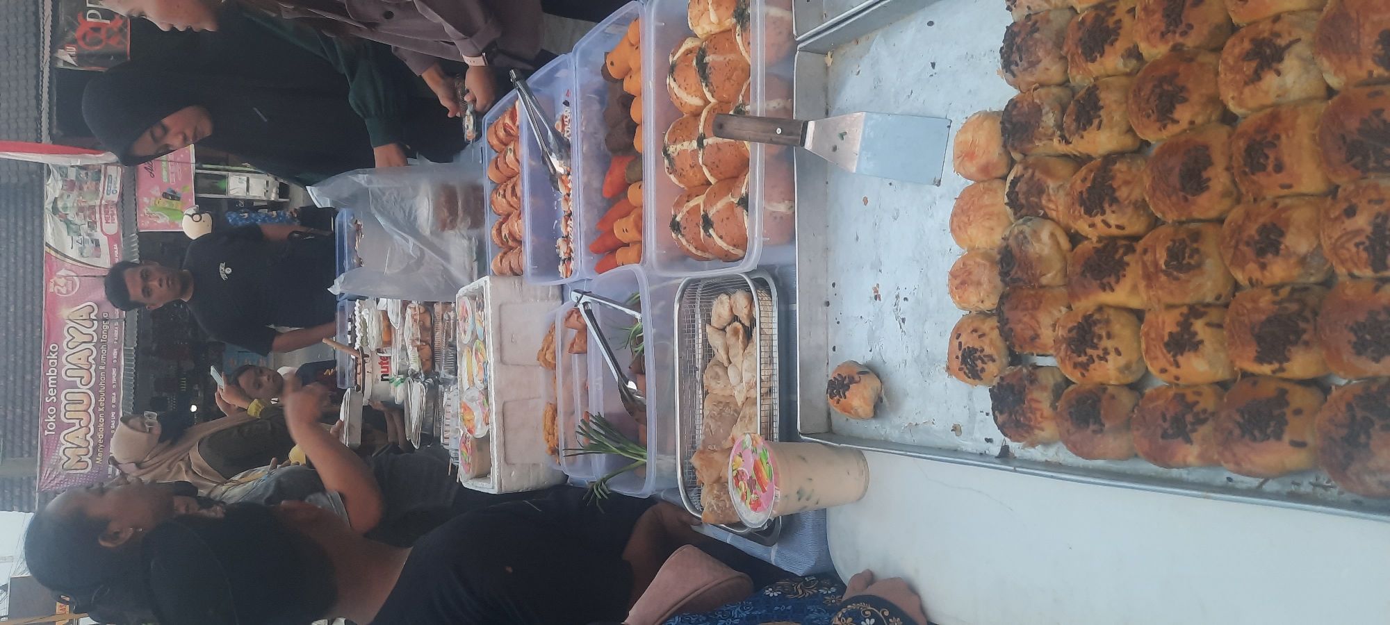 In Selopuro, Indonesia, a street food vendor displays a variety of pastries and snacks. A woman in a black hijab and a person with a brown hijab are visible on the right, attending to customers. In the center, a man in a black t-shirt stands behind a stall that shows the "MAJU JAYA" logo, indicating a "Toko Sembako" or grocery store. The foreground features trays filled with baked goods, some topped with chocolate sprinkles and cheese. To the left, a cup of a beverage with a colorful lid sits next to a tray of fried snacks. In the background, other individuals are seen browsing and purchasing items, suggesting a busy market atmosphere. The scene is set outdoors, likely on a street or sidewalk, with the presence of food stalls and people engaged in commerce.