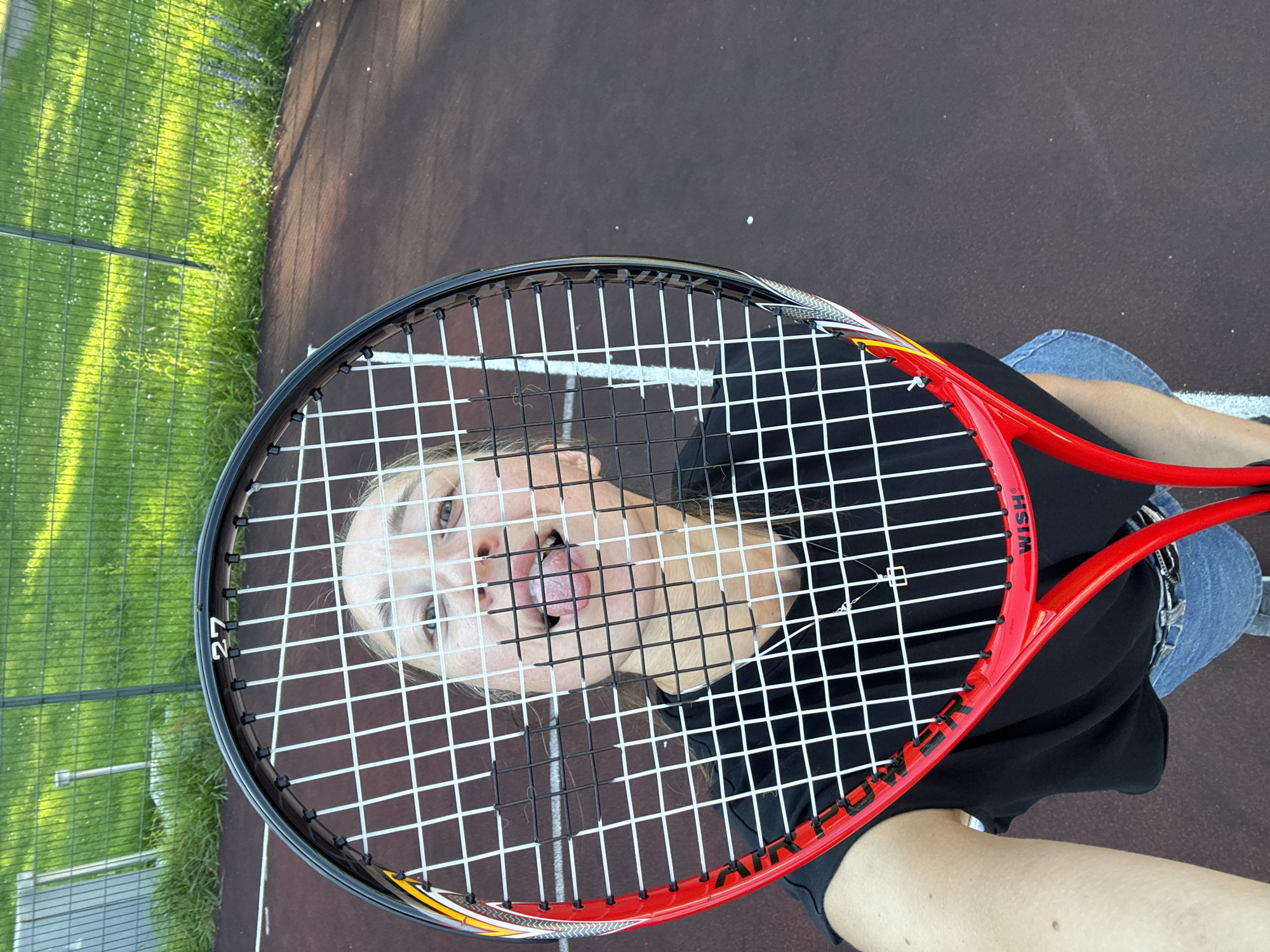 A young woman with blonde hair is taking a selfie on a tennis court, holding a tennis racket in front of her face. She is sticking her tongue out playfully through the strings of the racket. She is wearing a black t-shirt and blue denim shorts. The tennis court is brown with white lines and a green fence and grass are visible in the background. The image appears to be taken in the city of Perm, Russia, as indicated by the term "Прикамский" which is a geographical region in Perm Krai.