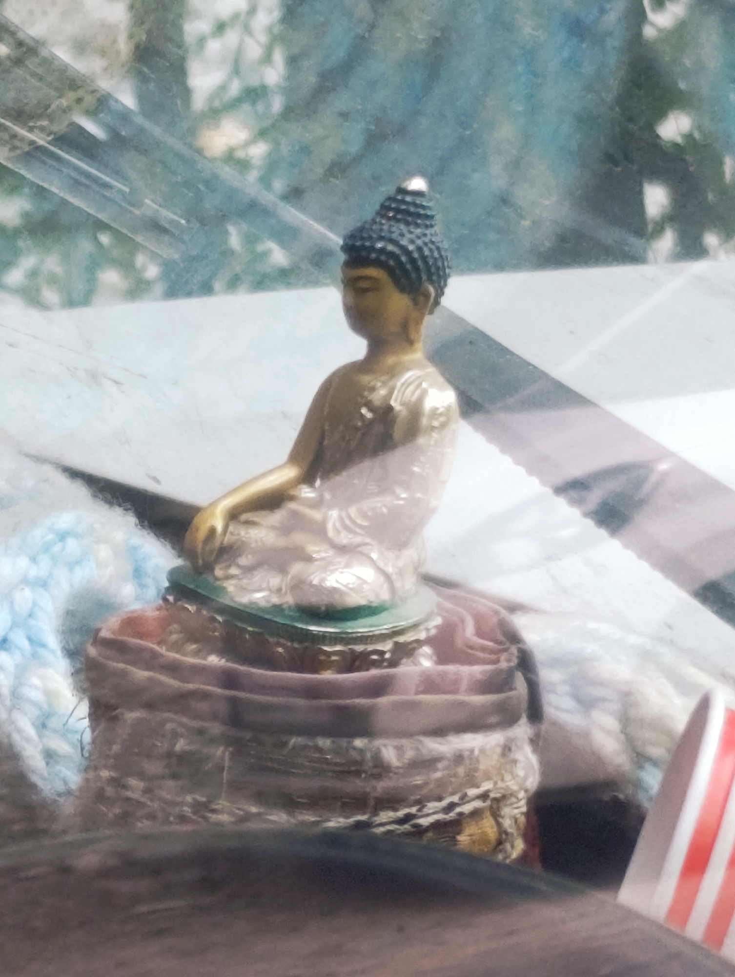 A small, metallic statue of Buddha is seated in a meditation pose on a multi-layered, fabric-covered base. The statue is gold-colored with dark accents on its robe and hair. The base is composed of circular layers of fabric in shades of brown and pink. To the left of the base, a fluffy, light blue and white material is visible. The background is blurred and appears to be a mix of green foliage and a reflective surface, possibly glass or a window. A portion of a striped object, red and white, is visible on the far right of the image. The scene is indoors, with sunlight suggesting it is daytime. The location is identified as Vashisht, India.