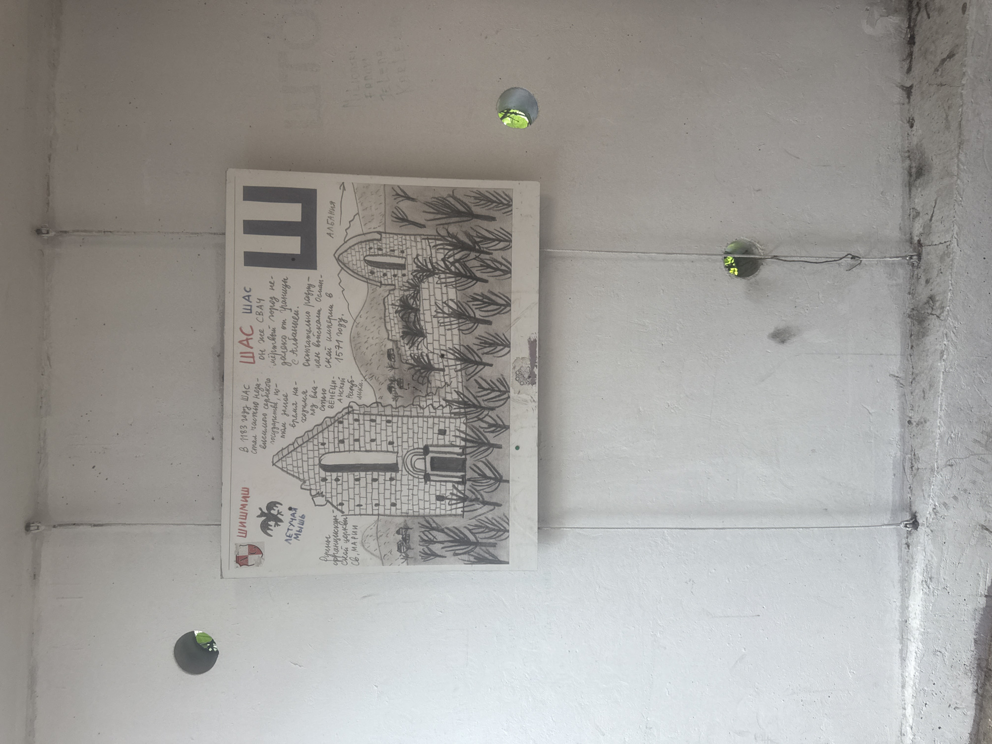 This is a photograph of a poster attached to a concrete wall, likely in an outdoor or semi-outdoor setting. The poster is oriented vertically and features a drawing of a stone structure, possibly a castle or church, surrounded by stylized trees. The drawing is in black and white with some text written in what appears to be Cyrillic and Latin script.
The Cyrillic text on the poster, translated, describes the historical site "Šaš Šaš" (also known as Svač), a "dead city" near the border with Albania. It details its history, noting it became part of an independent Serbian state in 1183, was later under Venetian rule, and was ultimately destroyed by the Ottoman Empire in 1571. The text also mentions "Šišmiš" (which translates to "bat") and "Letećaja Miš" ("flying mouse") along with an emblem of a bat and a shield. Another section of text appears to refer to a "Franciscan church of St. Mary." The Latin script "ALBANIJA" is visible on the top right of the drawing.
The concrete wall shows signs of wear and tear, with some staining and rough textures. Several thin wires are stretched across the wall, holding the poster in place. There are also three circular holes in the wall, two of which reveal a glimpse of green foliage, suggesting the presence of trees or plants outside. The lighting appears to be natural, casting shadows and indicating daylight. The overall impression is one of historical documentation or art displayed in an urban or industrial environment.