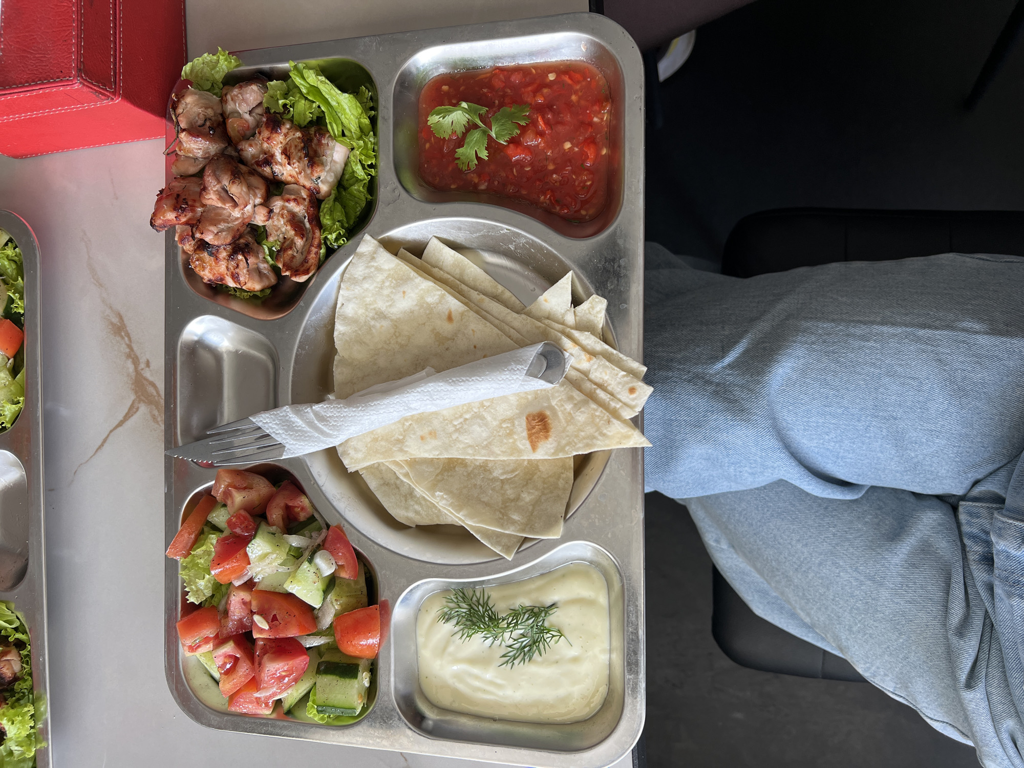A high-angle shot captures a metal tray divided into compartments containing various food items. The tray is positioned next to a person wearing light blue jeans. The food items include grilled meat on lettuce, a red sauce garnished with cilantro, tortilla wraps, a salad of tomatoes and cucumbers, and a creamy sauce with dill. A fork and knife wrapped in a napkin are on the tray. A red box is partially visible in the upper left corner. The scene appears to be indoors. It is impossible to determine the exact location of the city of North Kuta, Indonesia based solely on the image.