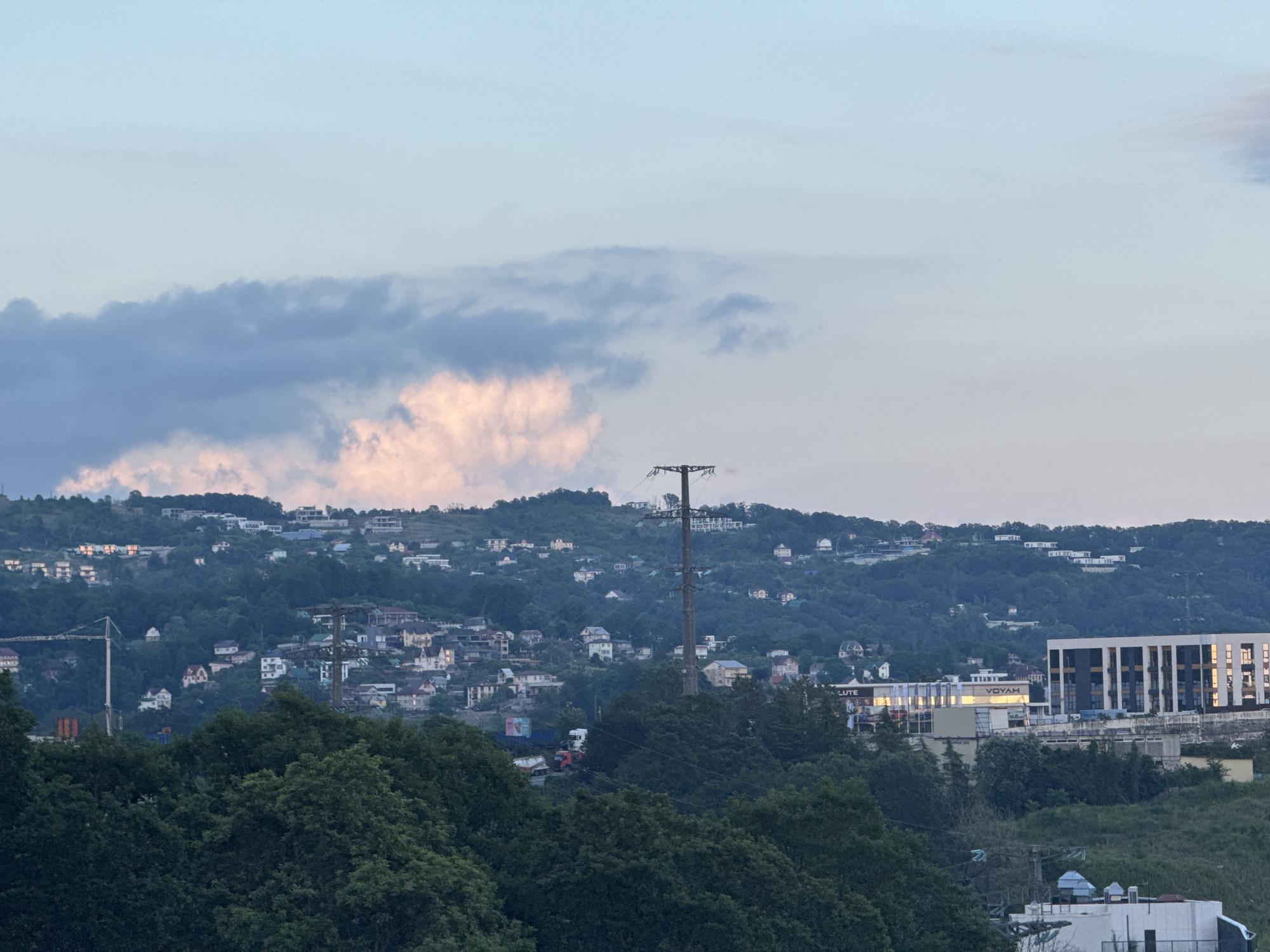 A wide-angle, eye-level shot captures a panoramic view of a coastal city nestled amongst rolling green hills under a pale blue and pink sky with scattered clouds. The foreground is dominated by dense, dark green foliage, suggesting a lush natural environment.
In the midground, the hillside is dotted with numerous residential buildings, varying in size and architectural style, many with visible windows reflecting the soft light of dusk. A prominent high-voltage electricity pylon stands tall on the hillside, a stark industrial element against the natural landscape. Further to the right, a modern, multi-story building with a distinctive facade of dark vertical lines and illuminated windows is visible, suggesting commercial or residential development. Smaller buildings and some infrastructure, possibly under construction or industrial, are also present.
In the background, the sky transitions from a soft pink near the horizon to a pale blue overhead, with some dramatic, light-colored clouds adding texture and depth to the scene. The overall impression is one of a city integrated into its natural surroundings, with a mix of residential development and modern infrastructure against a backdrop of hills and a serene sky. Based on the architectural styles and the hilly coastal terrain, this scene is likely in the city of Sochi, Russia, known for its mountainous landscapes and Black Sea coast.