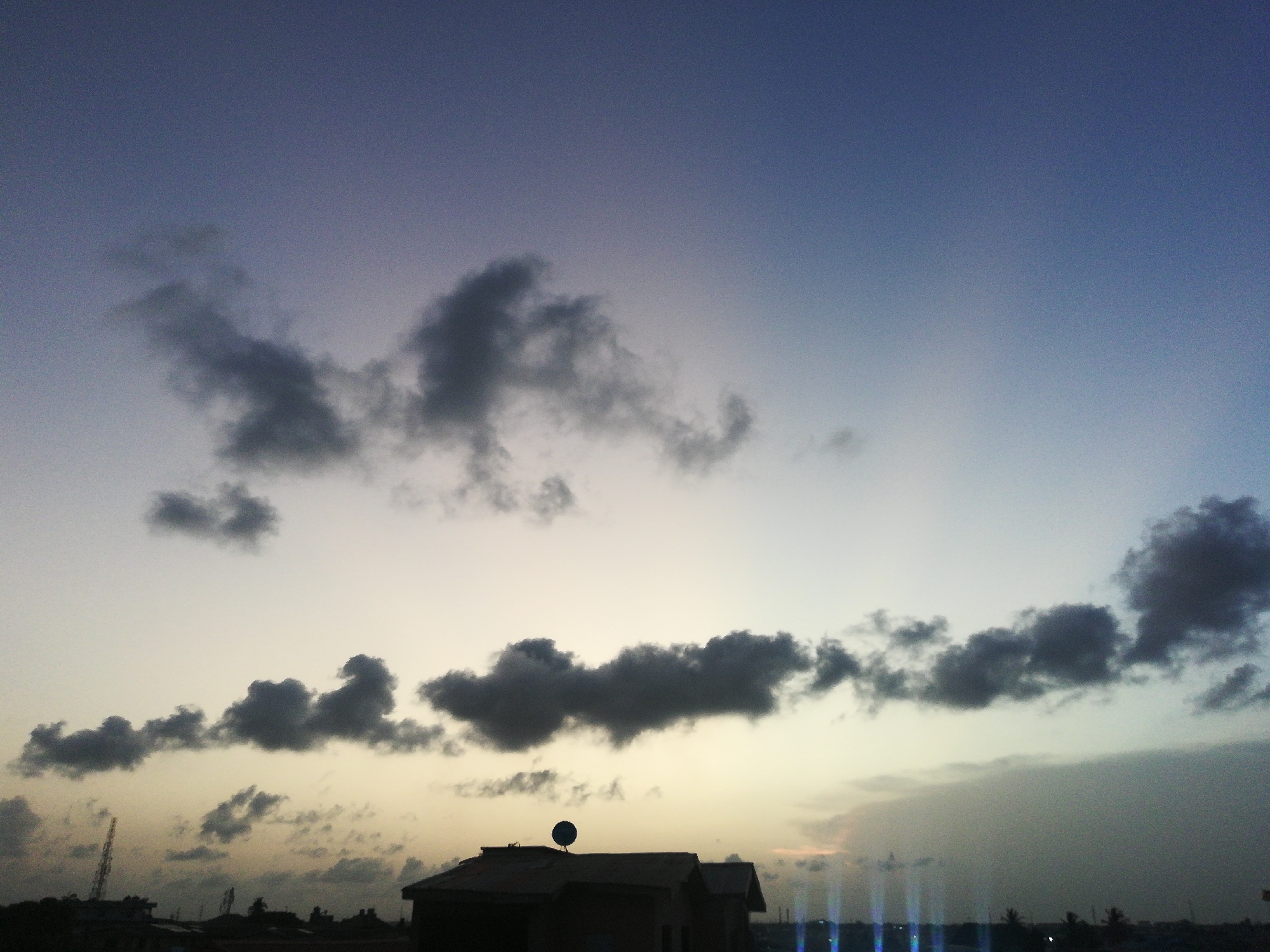 The image shows a twilight scene in Lagos, Nigeria. The sky transitions from a dark blue at the top to a light yellow-gray near the horizon. Several scattered gray clouds are visible. In the lower part of the image, silhouettes of buildings are seen, including a structure with a satellite dish on its roof. The skyline shows faint outlines of trees and possibly other buildings. Blue vertical streaks are present near the horizon, possibly indicating artificial light sources. No people are visible.