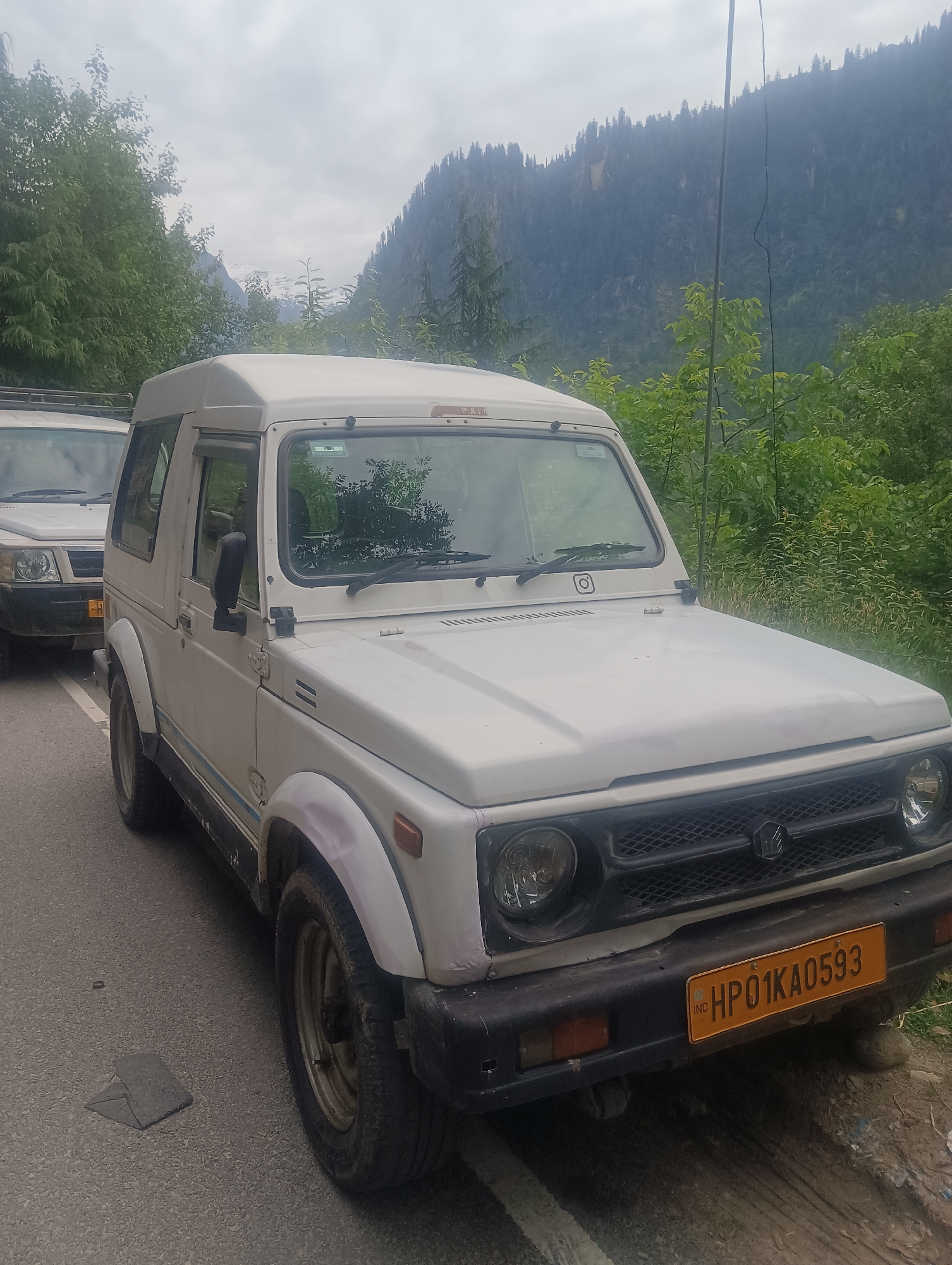 In Goshal, India, on a cloudy day, a white SUV is parked on the side of a paved road. The SUV has a black grille, round headlights, and a yellow license plate that reads "HP01KA0593". Behind the SUV, a mountain range covered in trees is visible. The weather is overcast.
