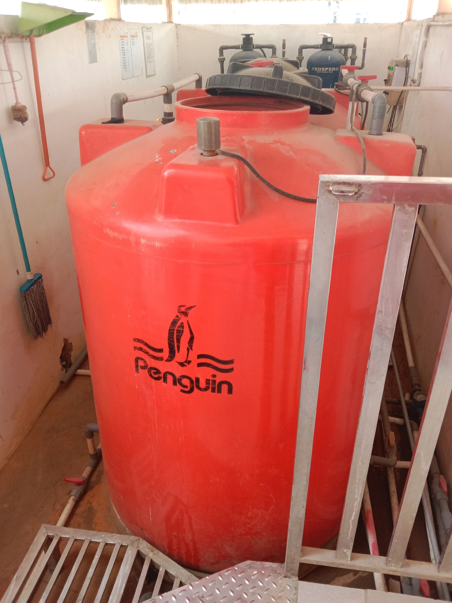 The image shows a large, red water tank with the "Penguin" logo on it. The tank is located in a room with white walls and a tiled floor. There are several pipes and valves connected to the tank, as well as some equipment labeled "PROSPERO". A broom and dustpan are leaning against the wall. The scene appears to be a water treatment or storage facility. The presence of the "Penguin" brand, a well-known Indonesian water tank manufacturer, and the general appearance of the facility suggest it could be located anywhere in Indonesia. Without further context or identifying markers, pinpointing the exact location within Semarang, Indonesia, is not possible based solely on this image.