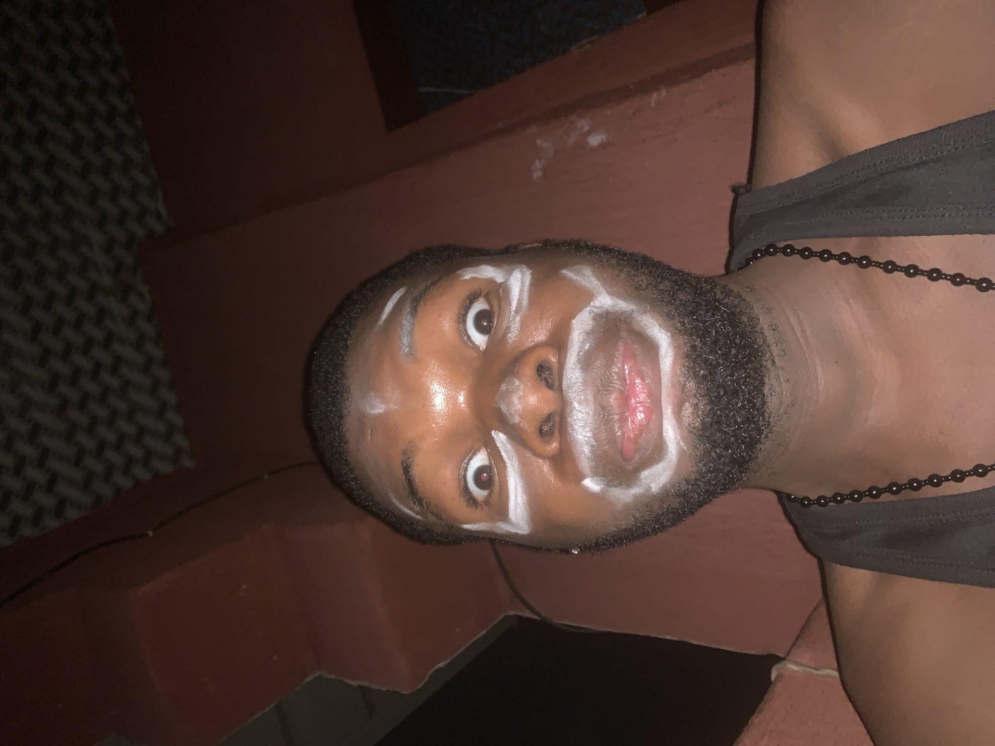 A close-up, rotated portrait shows a dark-skinned man with a short beard and hair, looking directly at the camera. White substance is applied to his face in distinct lines: above his eyebrows, along the bridge of his nose, and encircling his mouth, highlighting his features. His eyes are wide and focused, conveying an intense or perhaps startled expression. His mouth is slightly open, revealing red inner lips. He is wearing a dark, possibly sleeveless top, and a dark beaded necklace is visible around his neck. The background is dimly lit, consisting of dark reddish-brown structures that appear to be walls or steps, and a patterned surface, possibly tiled, on the left. The low lighting suggests a nighttime or indoor setting. The image, taken in Onitsha, Nigeria, captures a moment that could be part of a cultural ritual, performance, or personal artistic expression. No text is visible in the image.
