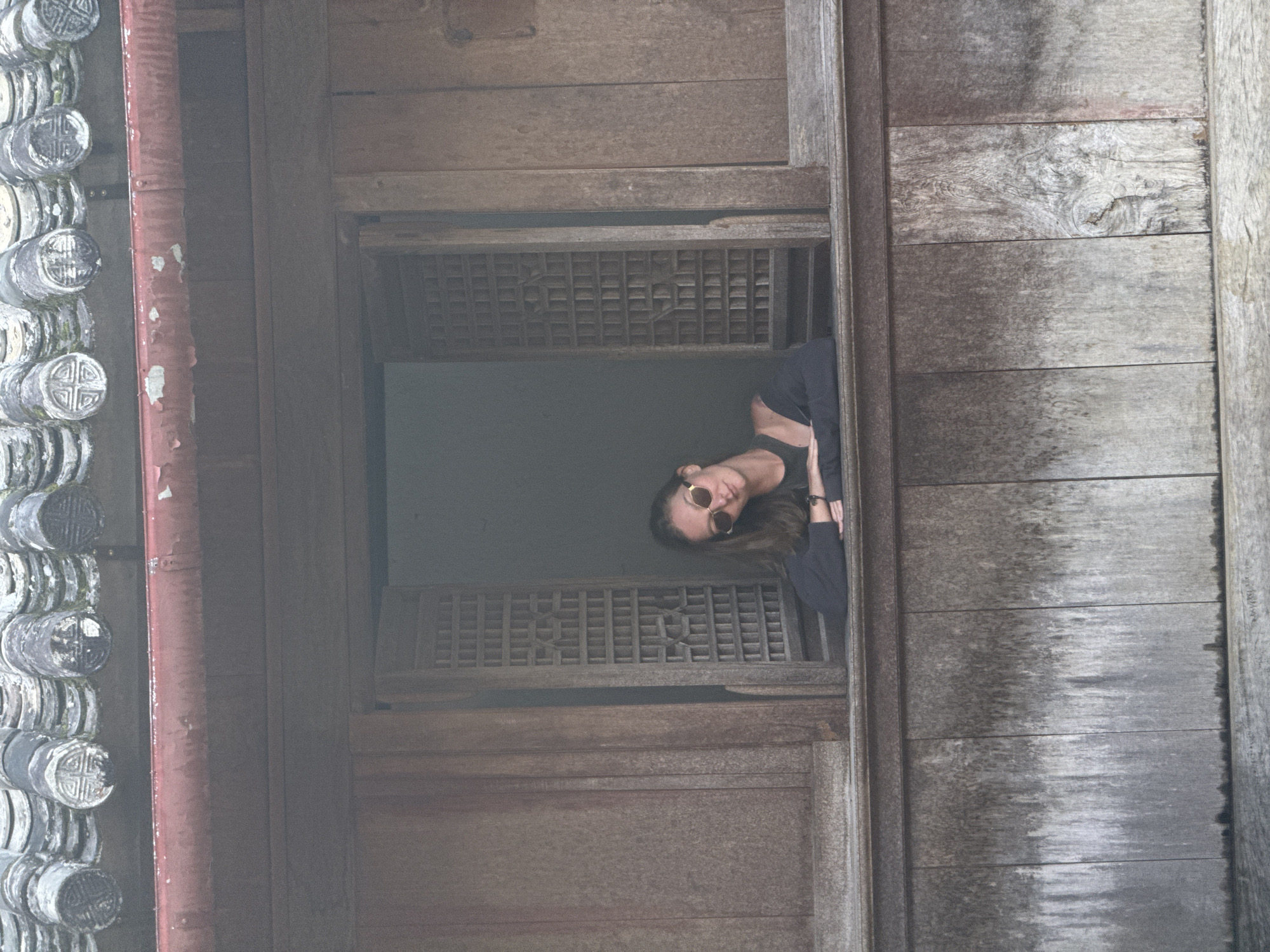 A rotated, medium shot shows a woman peering out of a wooden window in Sà Phìn Commune, Vietnam. The weathered window has a latticed screen. The woman is wearing sunglasses and a dark top. To the left of the window are traditional Vietnamese roof tiles. The wooden wall to the right of the window is aged and weathered.