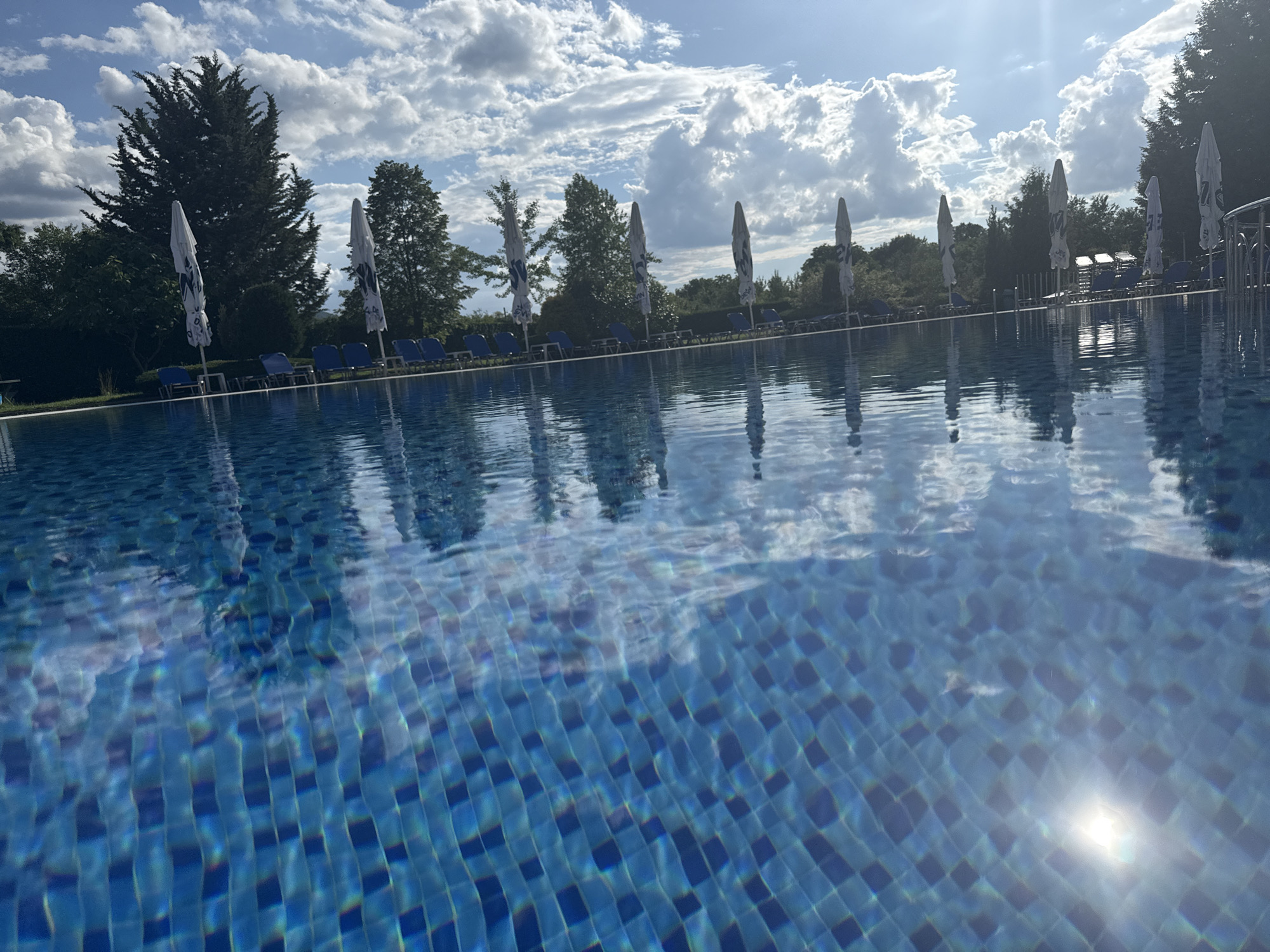 The image depicts an outdoor swimming pool with bright blue water tiled with a mosaic pattern. Sunlight reflects off the water's surface, creating a shimmering effect. In the background, several closed white umbrellas are positioned alongside rows of empty blue lounge chairs. Lush green trees and foliage border the pool area, and a bright, partly cloudy sky is visible above. The scene appears to be a resort or hotel swimming pool, likely in the city of Yagoda, Bulgaria, given the context. There are no people or animals present in the image.