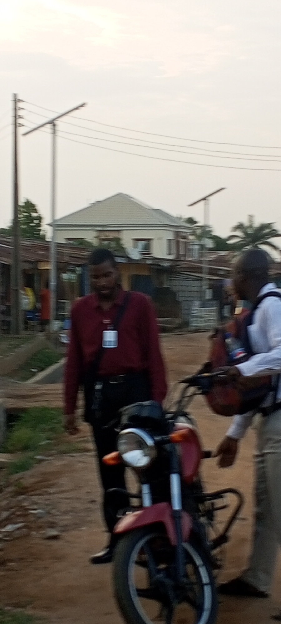 Two men stand by a red motorcycle on a dirt road in Sabon Tasha, Nigeria. One man wears a dark red shirt and dark pants and has an ID badge hanging from his neck. The other man wears a white shirt and light-colored pants and holds a red backpack. In the background, there are houses, utility poles, and power lines.