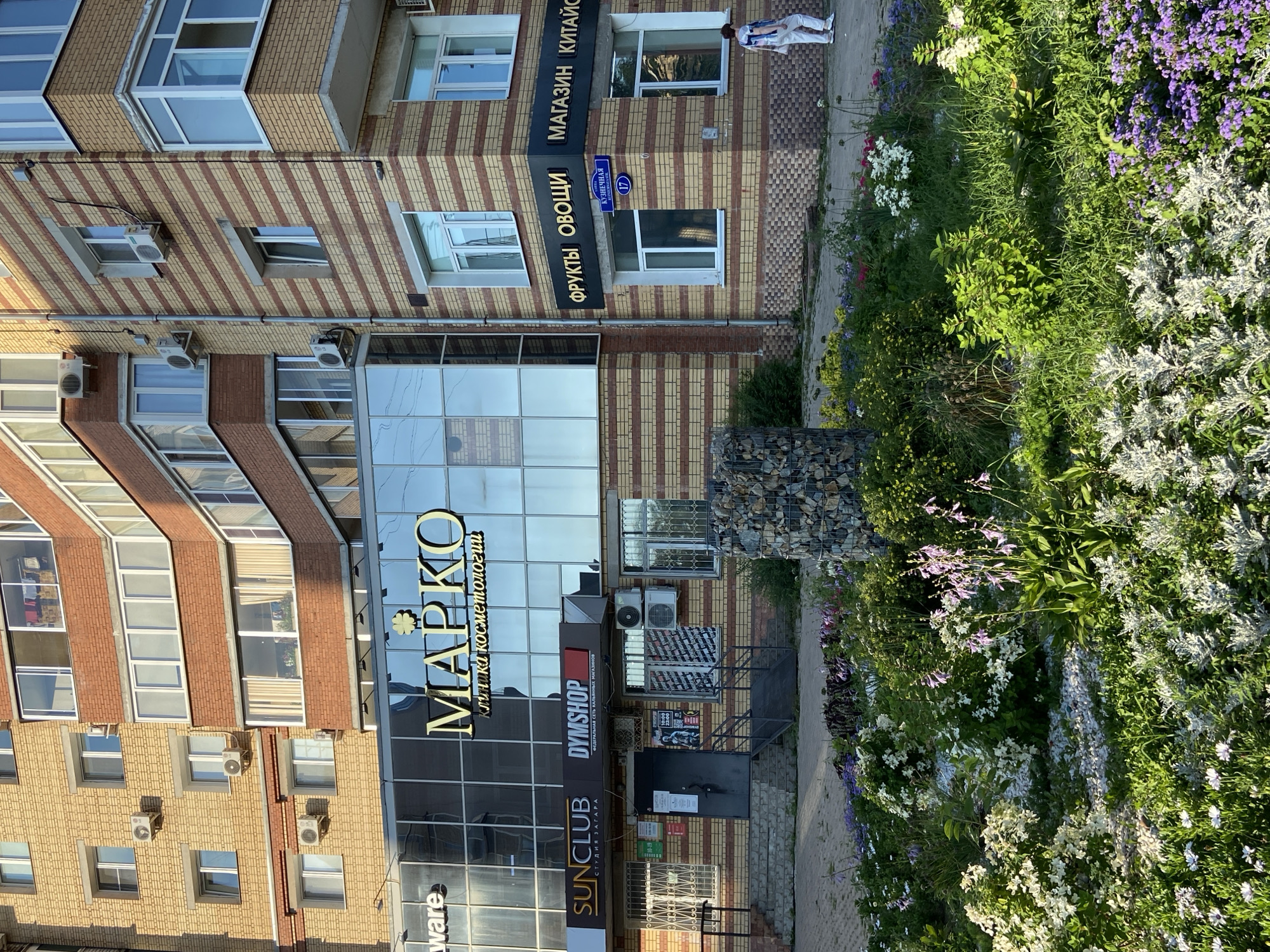 The image depicts a street-level view of a modern, multi-story building in Blagoveshchensk, Russia, with a focus on commercial establishments on the ground floor and a landscaped flower bed in the foreground.
The building's facade is a combination of light yellow bricks with horizontal darker brown bands and sections of brickwork. The upper floors feature numerous windows and balconies with white frames. Air conditioning units are visible mounted on the exterior walls.
The ground floor houses several businesses. A large glass storefront displays the prominent golden sign "МАРКО" with smaller text below indicating "клиника косметологии" (cosmetology clinic). Adjacent to this is a sign for "SUNCLUB" with the tagline "СТУДИЯ ЗАГАРА" (tanning studio), and a sign for "DYMSHOP" with the text "ФЕДЕРАЛЬНАЯ СЕТЬ КАЛЬЯННЫХ МАГАЗИНОВ" (federal chain of hookah stores). The entrance to the building is a dark door with stairs leading up to it. Window displays and posters are visible.
A street sign indicating "КУЗНЕЧНАЯ" and the number "17" is mounted on the building corner. Another sign with a blue circle and the number "17" is also visible.
In the foreground, a well-maintained flower bed features a variety of plants, including white and purple flowers, green foliage, and ornamental grass. A decorative cylindrical structure made of stacked stones is placed within the flower bed. The ground surface is paved with gray stones.
A person dressed in white pants and a light-colored top is walking away from the camera on the sidewalk to the right of the building. The lighting suggests it is daytime, with shadows cast on the ground. The overall scene conveys a sense of urban commercial activity integrated with green landscaping.