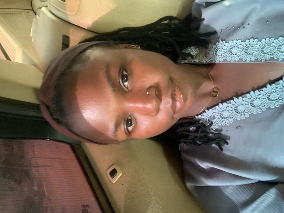 A young woman with dark skin and dark hair styled in dreadlocks, partially covered by a dark headband, is depicted in a close-up photograph taken from within a vehicle. She has a small, light-colored stud piercing in her right nostril and a gold-colored, elongated oval earring in her right ear. Her lips are glossy. She is wearing a light gray garment featuring an intricate white lace trim along the neckline and shoulders, accented with small, shiny embellishments. A gold-colored chain necklace with a "CD" pendant is visible on her chest. The background shows the light-colored interior of a vehicle, including a window on the left reflecting indistinct reddish-pink light, suggesting an external low-light or nighttime environment. A small, dark button is present on the interior panel adjacent to the window. The location is stated as Gadam, Nigeria, but this cannot be visually verified from the image content.