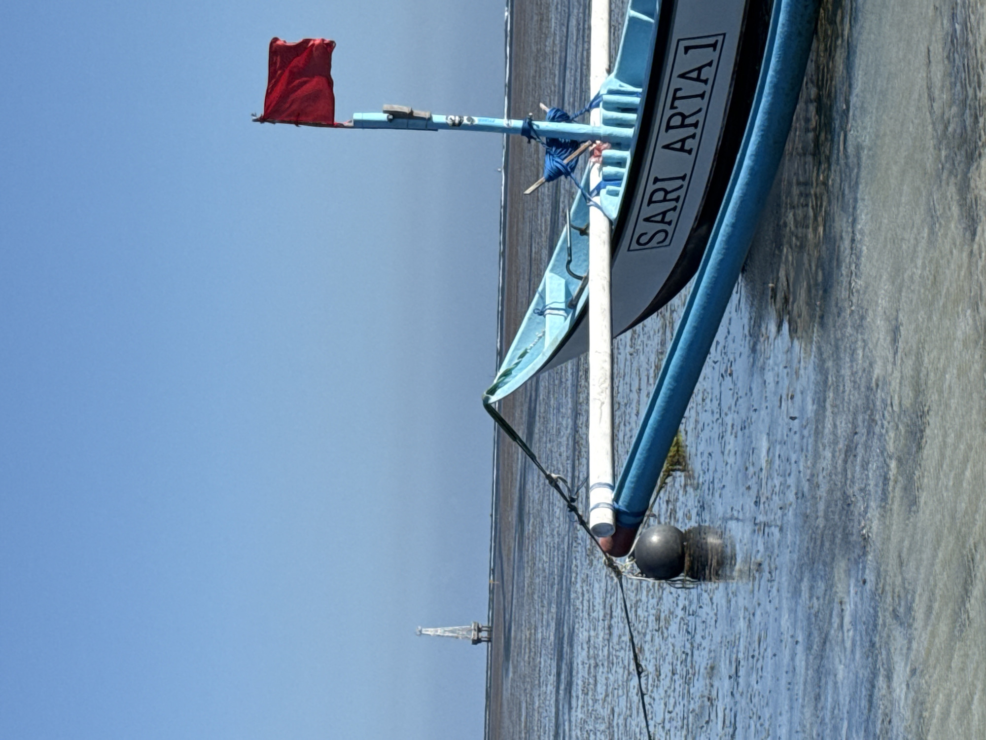 The image captures a fishing boat named "SARI ARTA 1" docked in shallow, calm water, possibly at low tide, near the coast of Denpasar, Indonesia. The boat, painted blue and white, has a red flag flying from its mast. In the distance, a white, slender structure, possibly a lighthouse or marker, stands on stilts in the water. The sky is a clear, bright blue. The water's surface reflects the sky and the boat, with visible sand or mud beneath the shallow water. A dark spherical buoy is tethered to the boat. The scene suggests a peaceful, coastal environment.