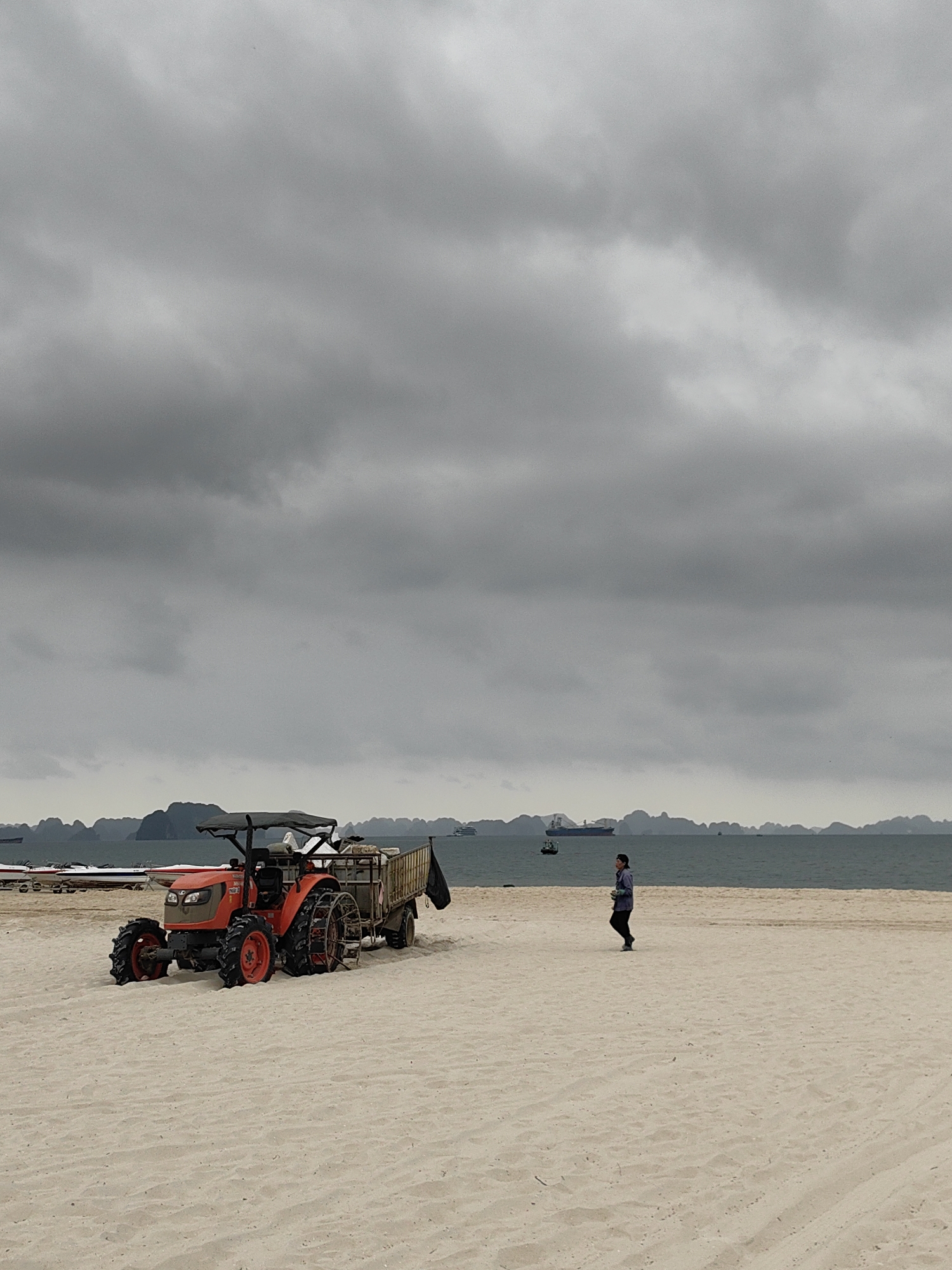The image shows a beach scene in Bai Chay Ward, Vietnam, under an overcast sky. A compact orange tractor with a trailer is parked on the sand near the left side of the frame. A person is walking on the beach in the distance to the right of the tractor. The sea is visible in the background with some boats and distant islands. The sand is pale and takes up the majority of the foreground.
