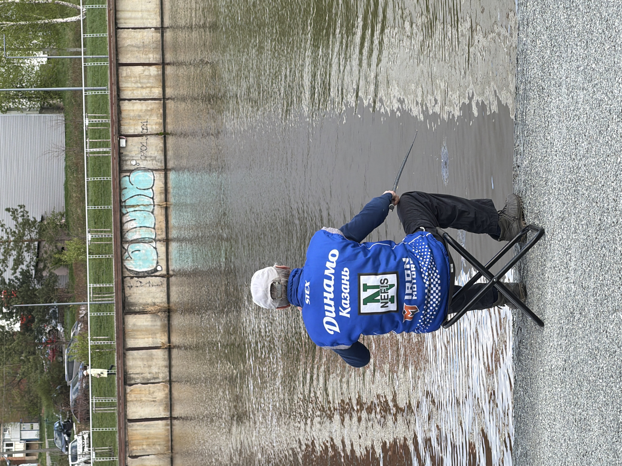 A man is fishing on the bank of the Votkinsk Reservoir in the city of Votkinsk, Russia. He is seated in a black folding chair on a gravel embankment. The man wears a white cap and a blue jacket with the words "Динамо Казань" and the logo "NEFIS". He holds a fishing rod over the water. Behind him is a concrete wall with some graffiti, and a metal ladder is affixed to the wall. Beyond the wall, there is a building, some trees, and parked cars.