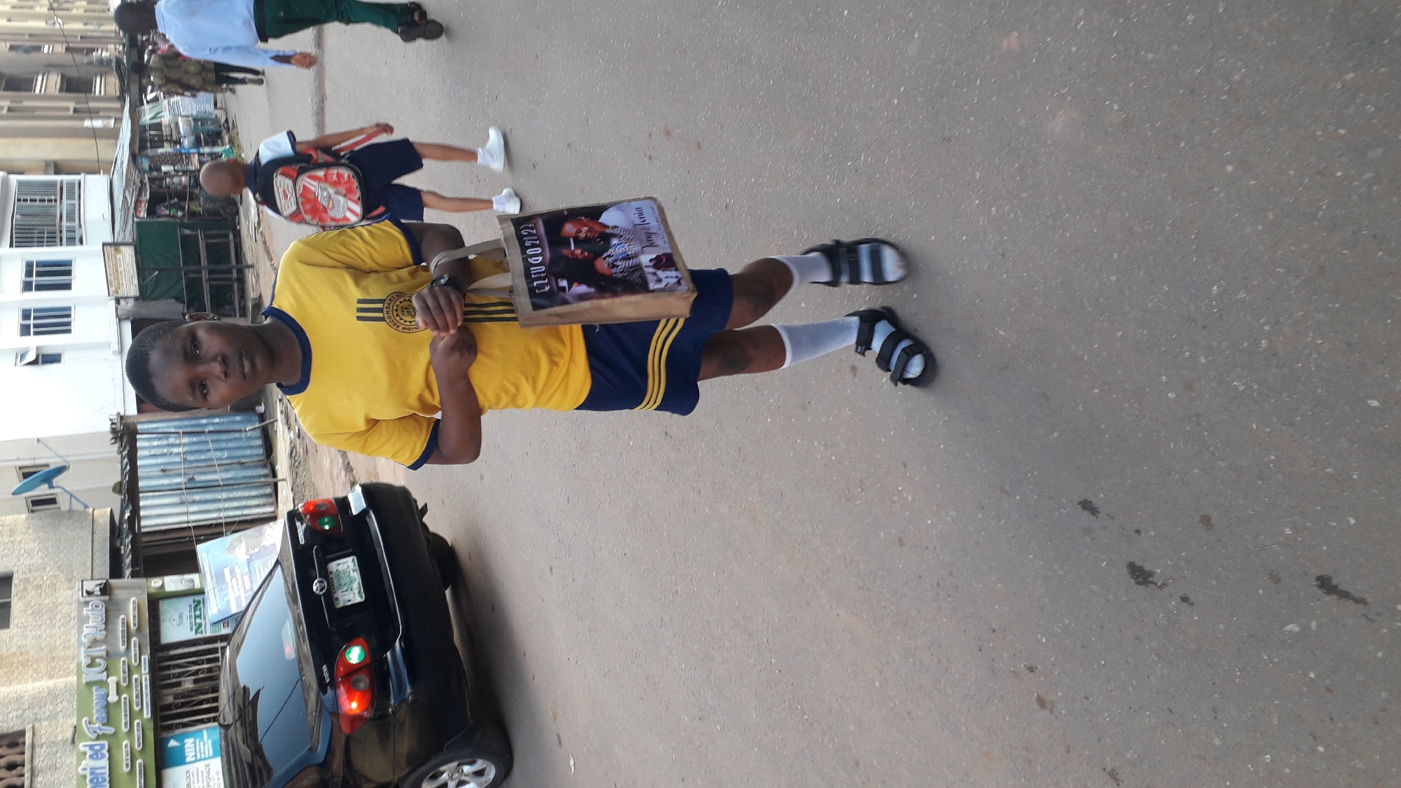 A young boy in a yellow and blue school uniform, white socks, and sandals stands on a paved road in what appears to be Onitsha, Nigeria, based on the visible signage. He's holding a tote bag. In the background are buildings, a car, and other individuals. A sign for "Merited Favor ICT Hub" is visible in the background along with a license plate.