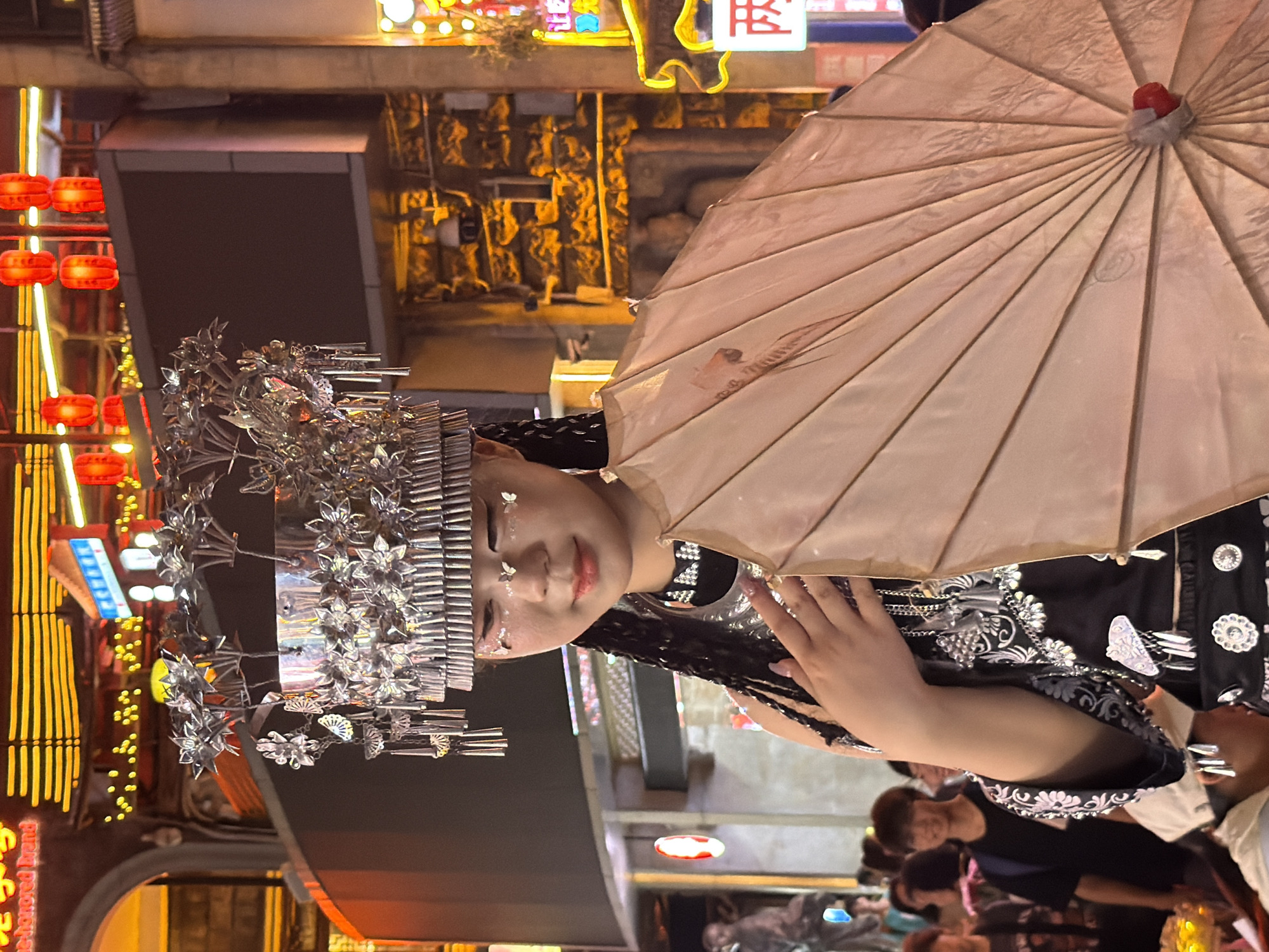 This media file captures a young woman dressed in traditional attire, possibly for a cultural event or performance. She wears an elaborate silver headdress adorned with floral motifs and intricate metalwork. Her outfit is a black garment with white floral embroidery and decorative silver buttons. She is holding a light-colored, possibly paper, umbrella, tilted to her side. The woman has glittery makeup on her face, including what appears to be small butterfly designs. She has long, dark braided hair.
The setting appears to be an outdoor night market or festival, indicated by the abundant colorful lights and illuminated signs in the background. Red lanterns are visible on the left, and a neon sign with Chinese characters is also present. The atmosphere seems festive and lively, with other people visible in the background, some of whom appear to be seated and watching. The overall impression is one of a vibrant cultural celebration. The time of day is clearly nighttime due to the artificial lighting and dark surroundings. The location is implied to be Yuzhong District, China.