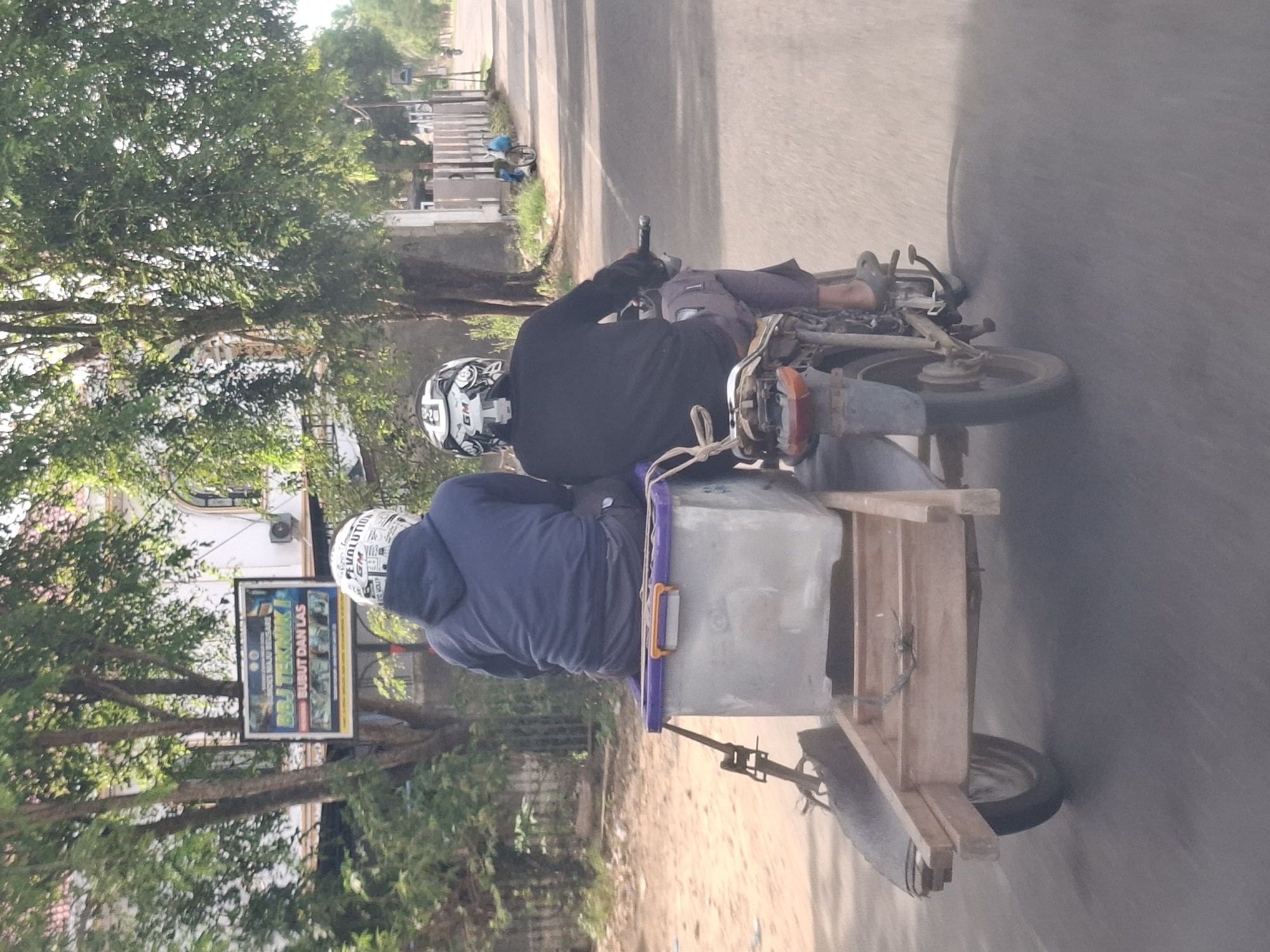 In Meuse, Indonesia, two individuals are riding a motorcycle with a trailer. The person in the front is wearing a blue hoodie and a white helmet with black and silver graphics. The person in the rear is wearing a black long-sleeved shirt and a black helmet with similar graphics. The motorcycle is towing a wooden trailer with a large, gray container with a purple lid. The trailer appears to be constructed from simple wooden planks and has one wheel. The scene is outdoors, with trees and buildings visible in the background. A sign with Indonesian text is mounted on a pole to the left of the road. The road is paved, and there is a dirt or gravel shoulder on the left side. The overall impression is a functional mode of transport for goods or supplies.