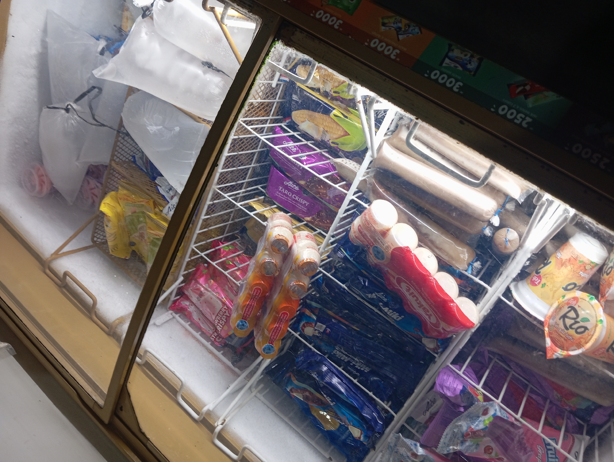 This is an eye-level, close-up shot of the interior of a chest freezer displaying various frozen food items and what appear to be ice packs. The freezer is divided into sections by white wire racks. In the foreground, to the left, are several colorful packages, including pink "Strawberry Crispy" items and blue and dark blue packages of ice cream or popsicles, some labeled "Milo". To the right of these, several orange and white packaged items are visible, possibly ice cream bars. Further back, there are purple packages of "Taro Crispy" and a package depicting corn on the cob. In the upper portion of the freezer, several large, translucent plastic bags filled with ice or frozen water are visible, along with smaller yellow-wrapped packages. The lighting is dim and casts shadows. On the right side of the image, part of a price list or advertisement with the numbers "2500" and "3000" printed on it is visible. The scene appears to be a retail freezer in a shop or market in Rejosari, Indonesia.